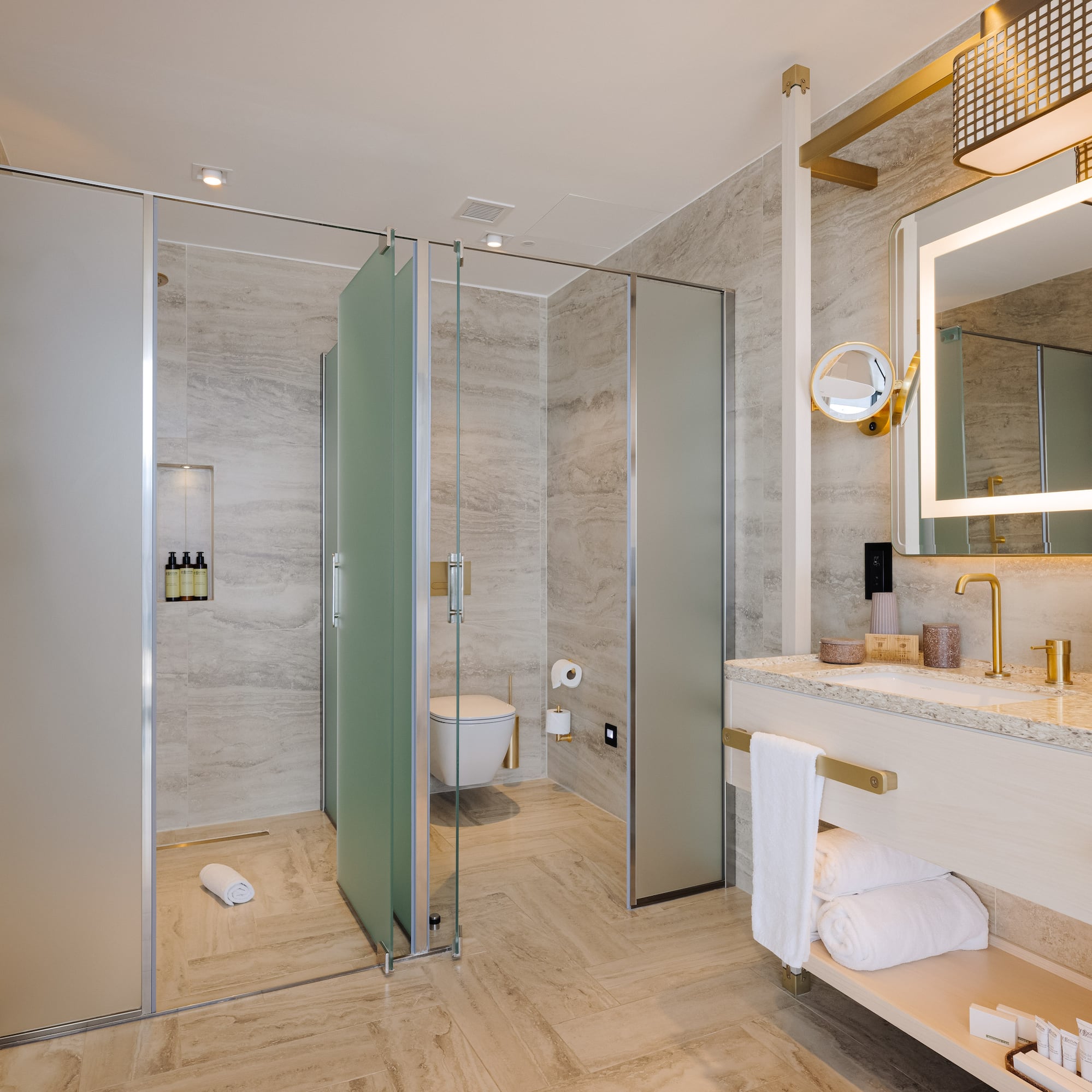 Modern luxury bathroom with gold fixtures, lighted mirror, and glass shower.