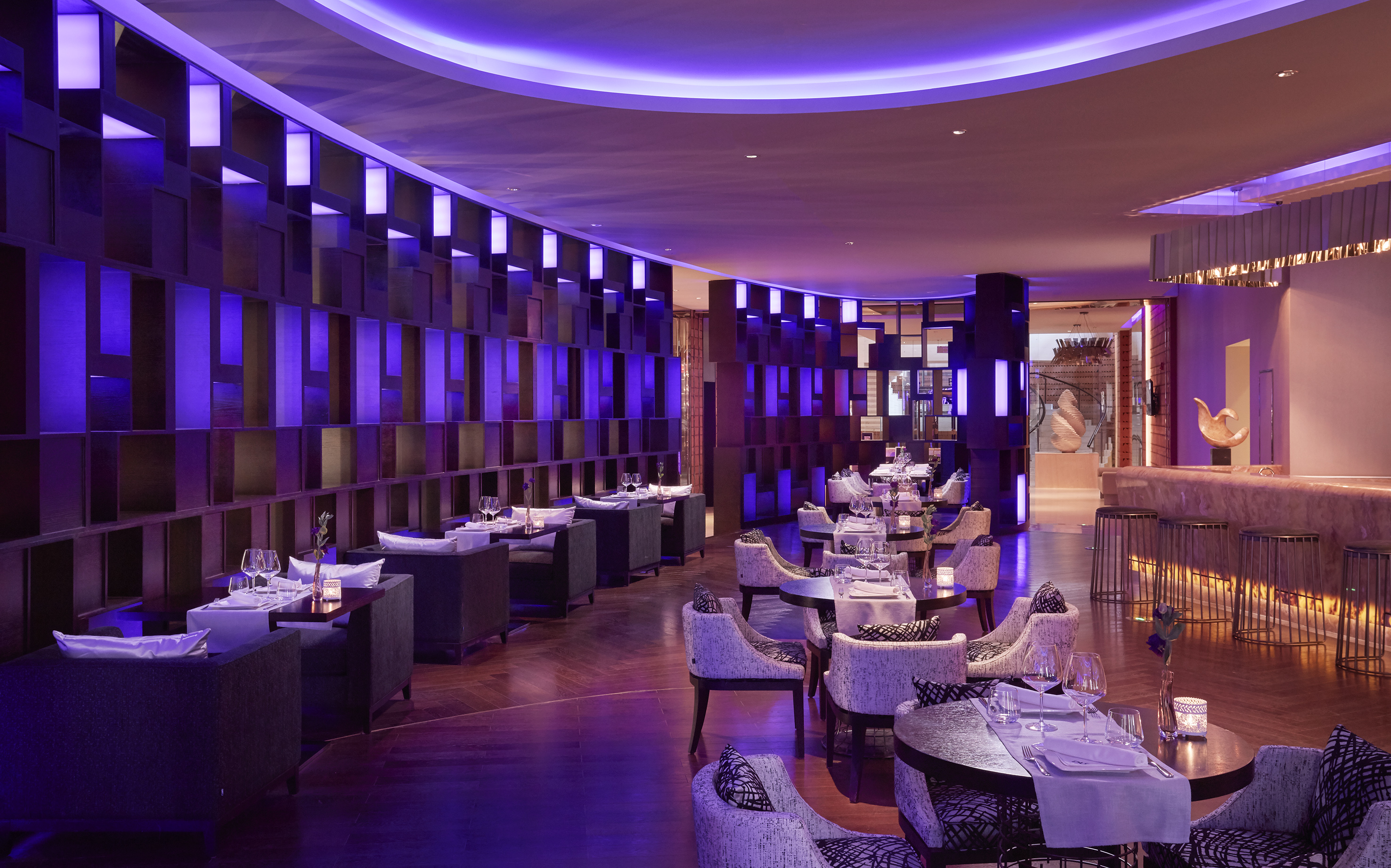 Upscale restaurant with purple lighting, wood floors, set tables, and modern bar.