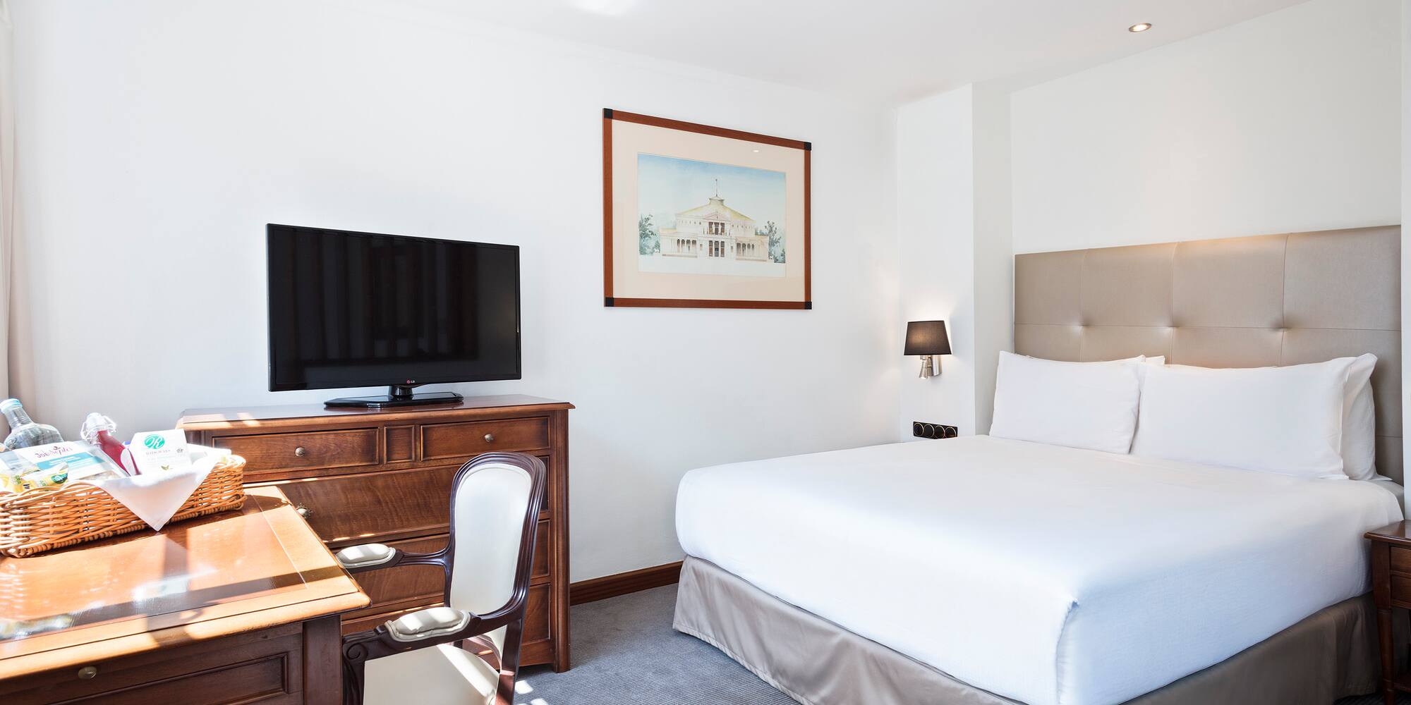 Bright hotel guest room with king bed, TV, wooden desk, and beige headboard.