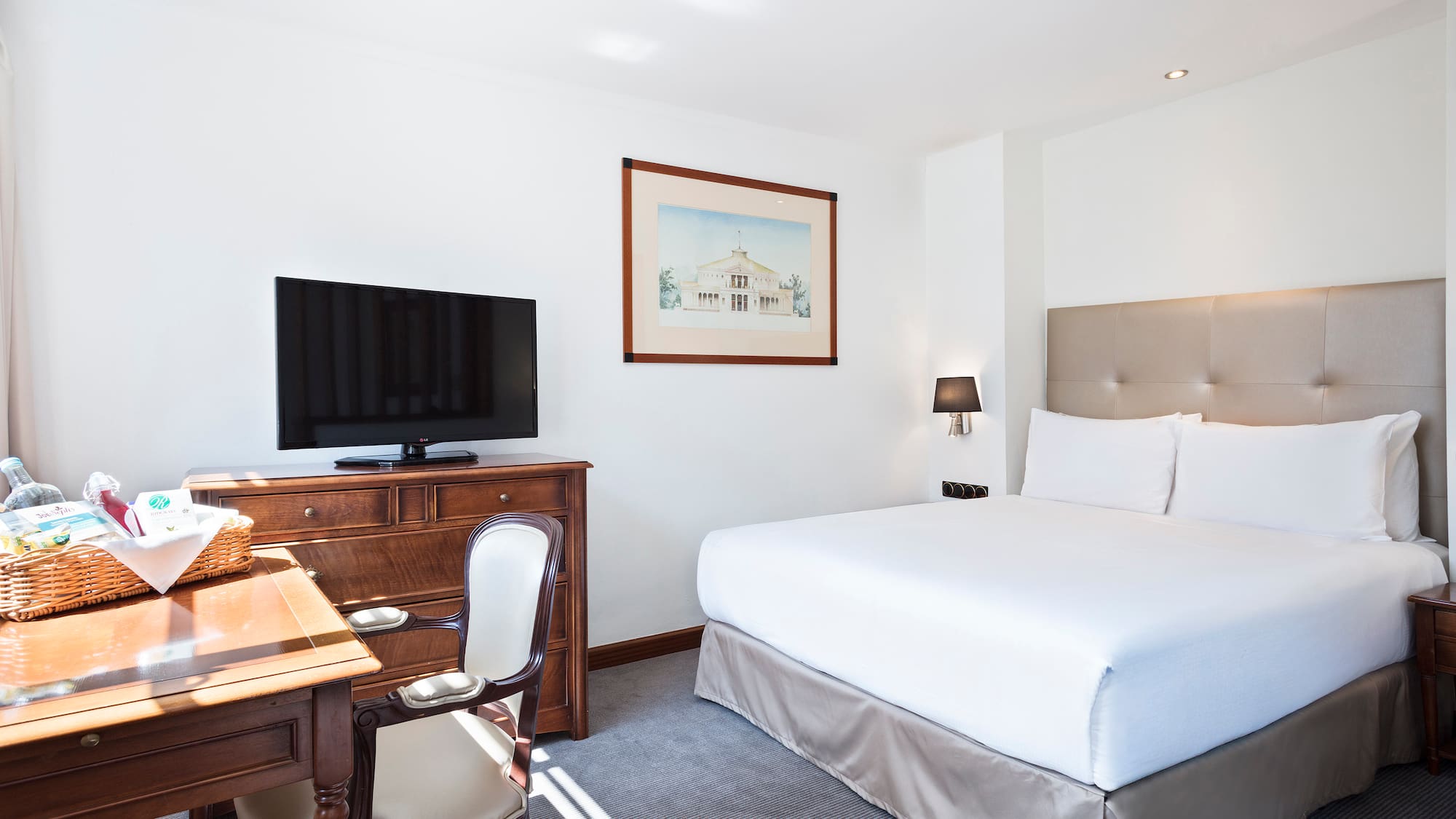 Bright hotel guest room with king bed, TV, wooden desk, and beige headboard.