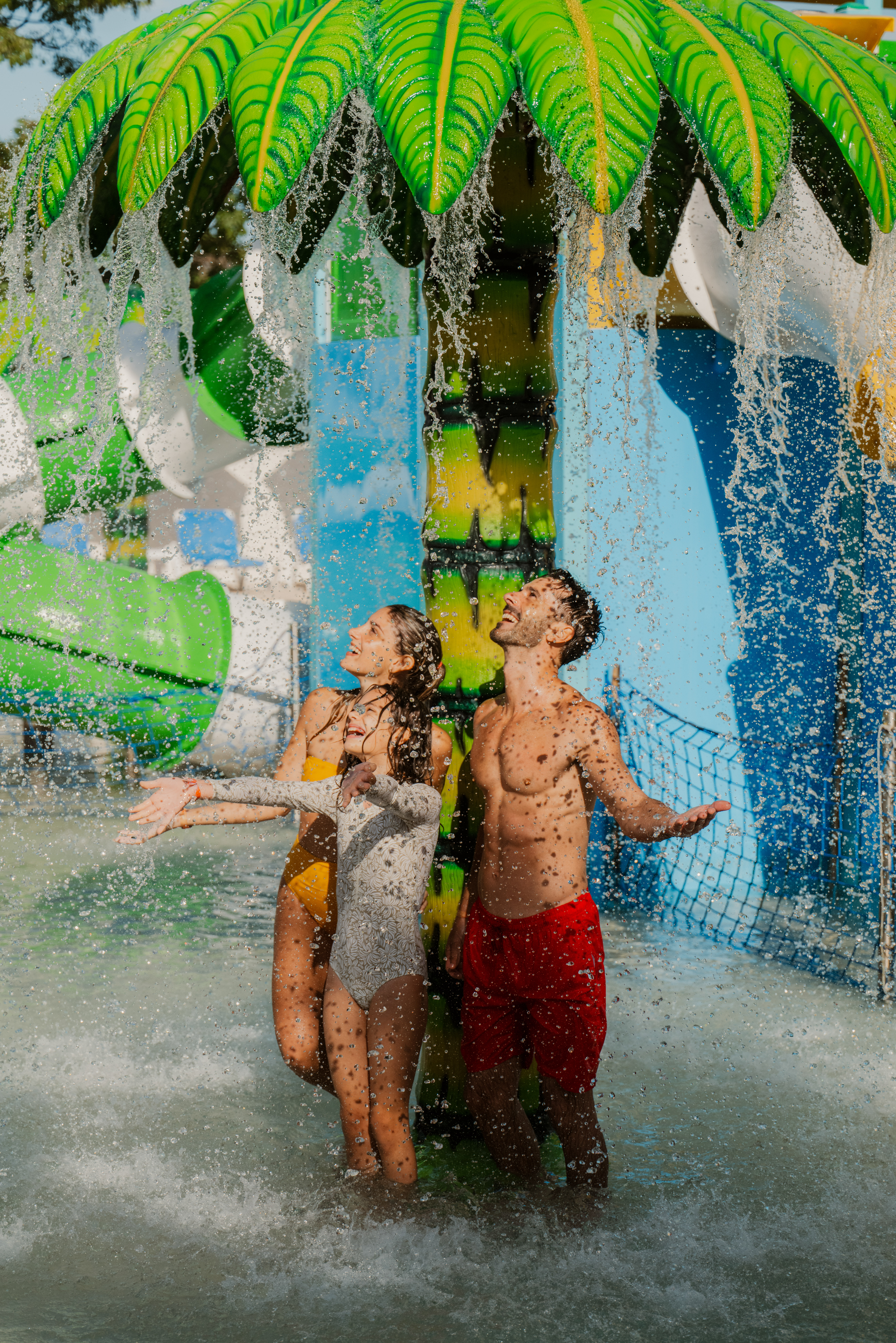 Joyful family playing under a palm tree water feature at a water park.