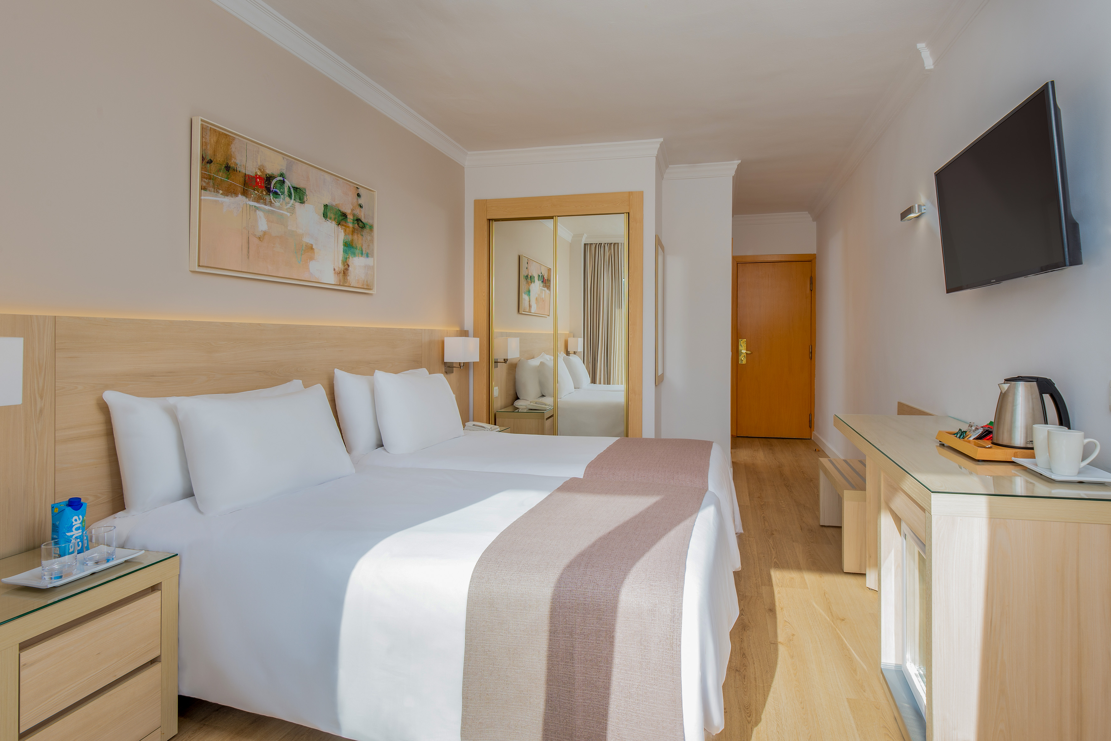 Modern hotel room with twin beds, mirrored closet, TV, and desk.