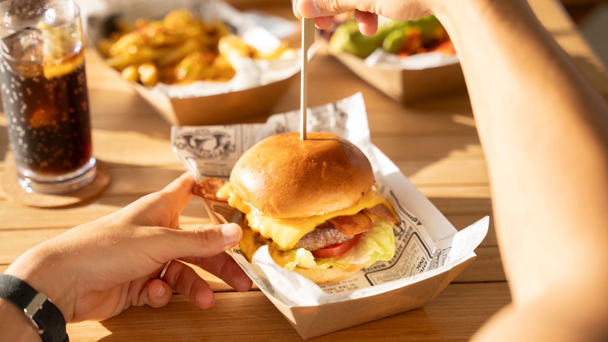 Hands holding a bacon cheeseburger with fries and soda on a wooden table.