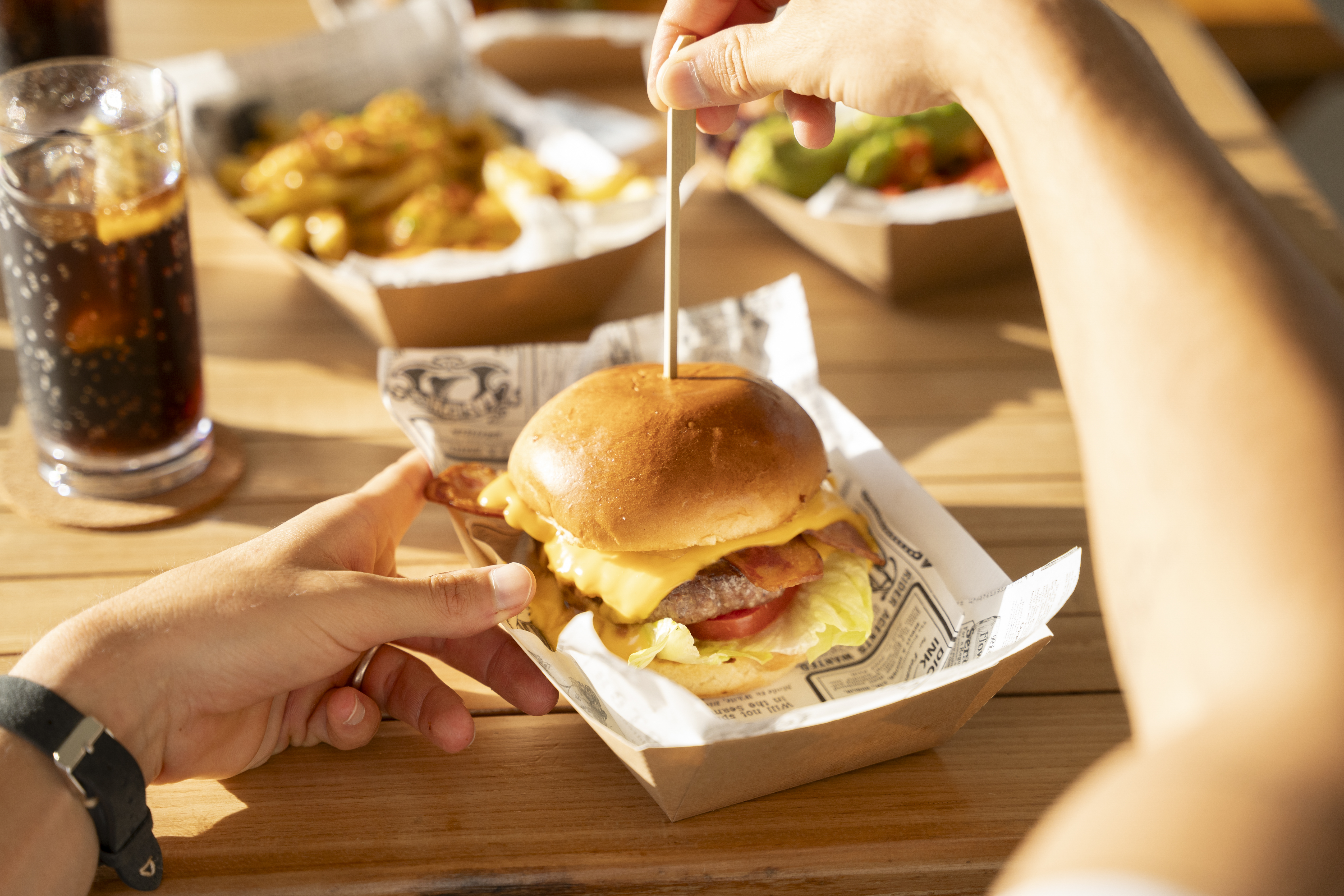 Hands holding a bacon cheeseburger with fries and soda on a wooden table.