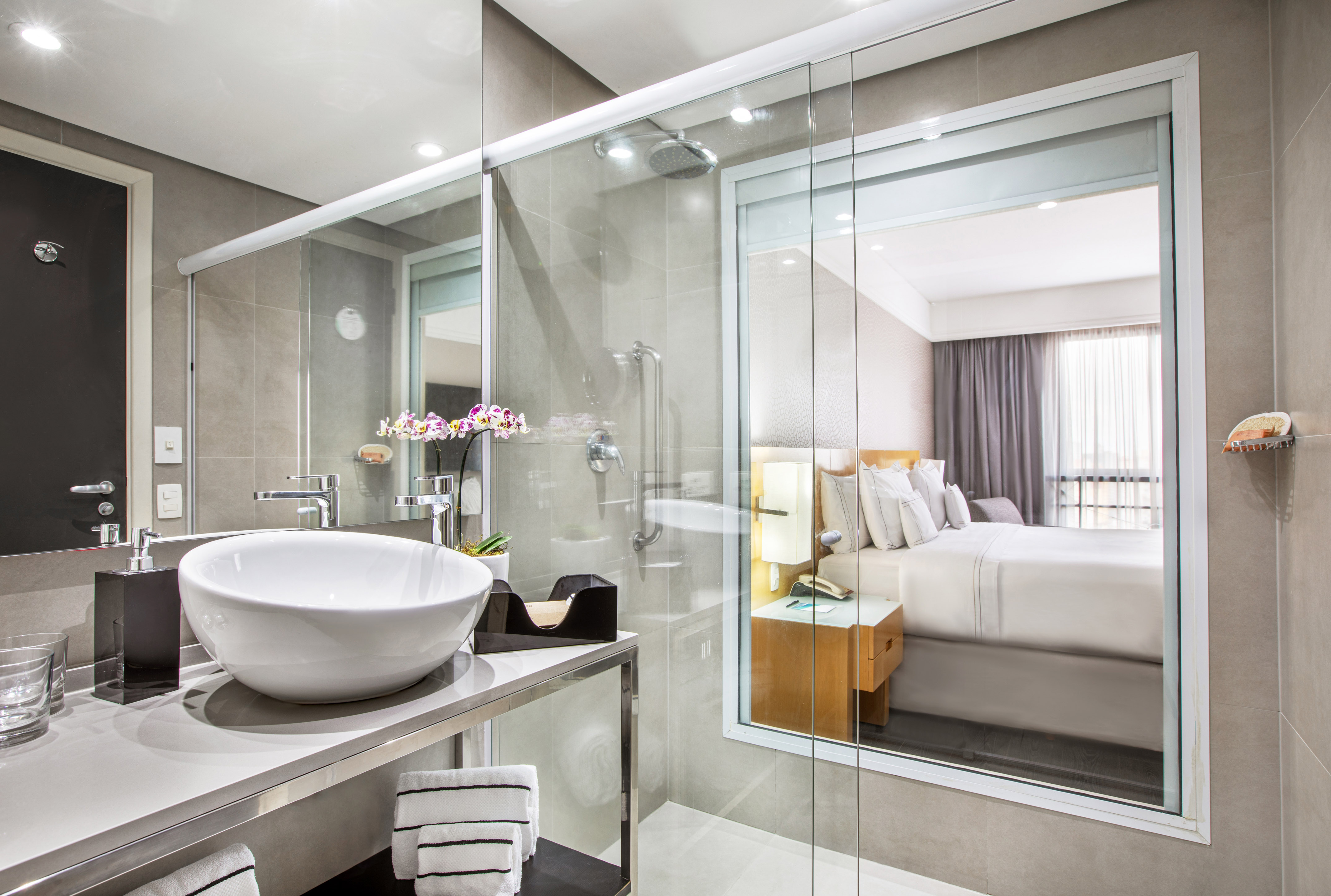 Modern hotel bathroom with vessel sink, glass shower, and bedroom view.