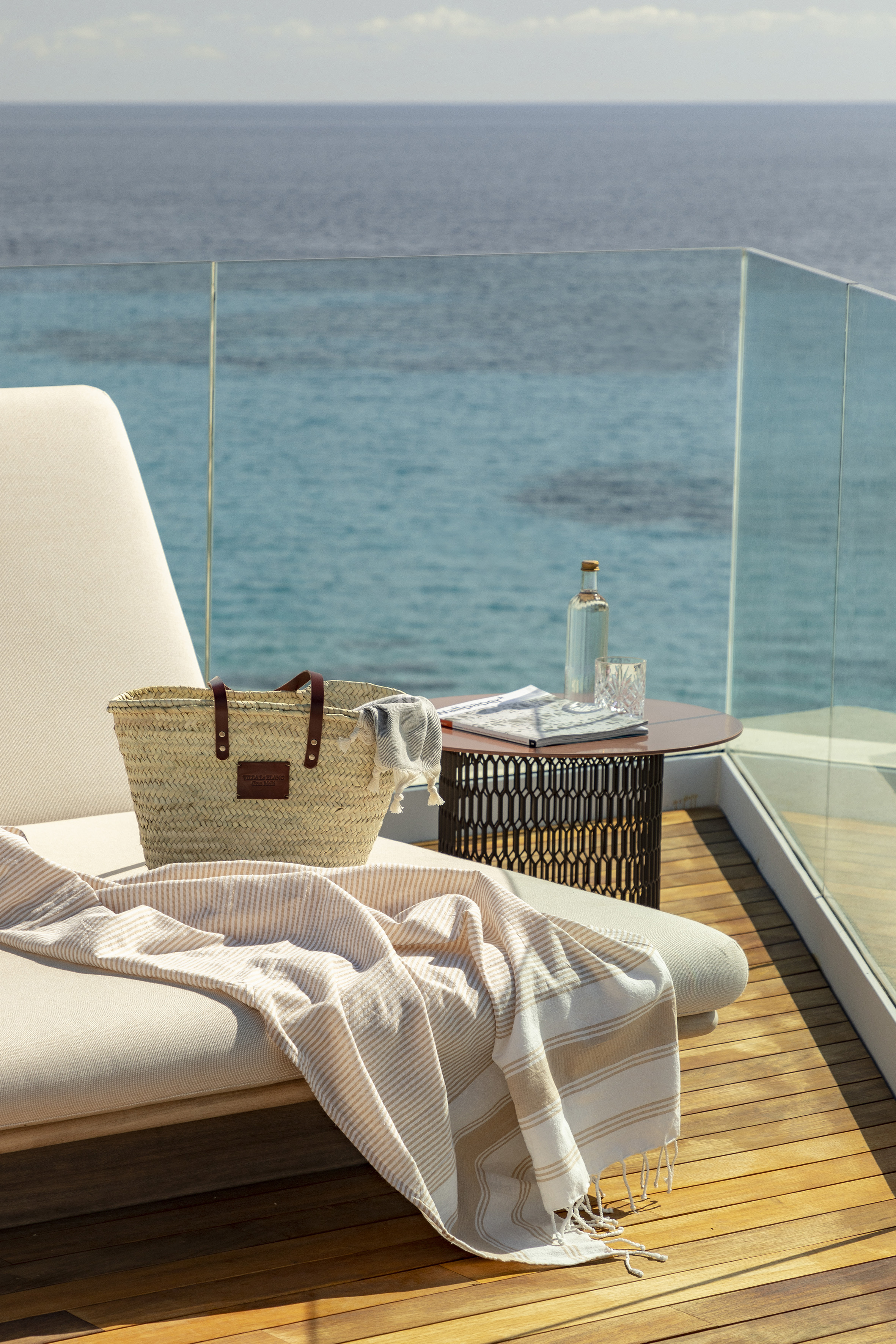Oceanfront balcony with sunbed, straw bag, and striped beach towel by the sea.
