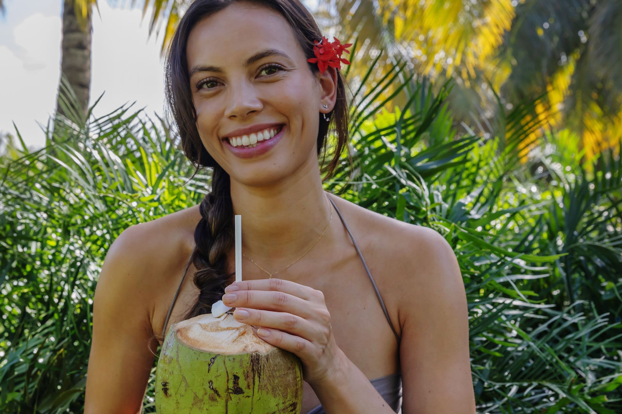 Mujer sonriendo, sosteniendo coco fresco con popote entre palmas tropicales.