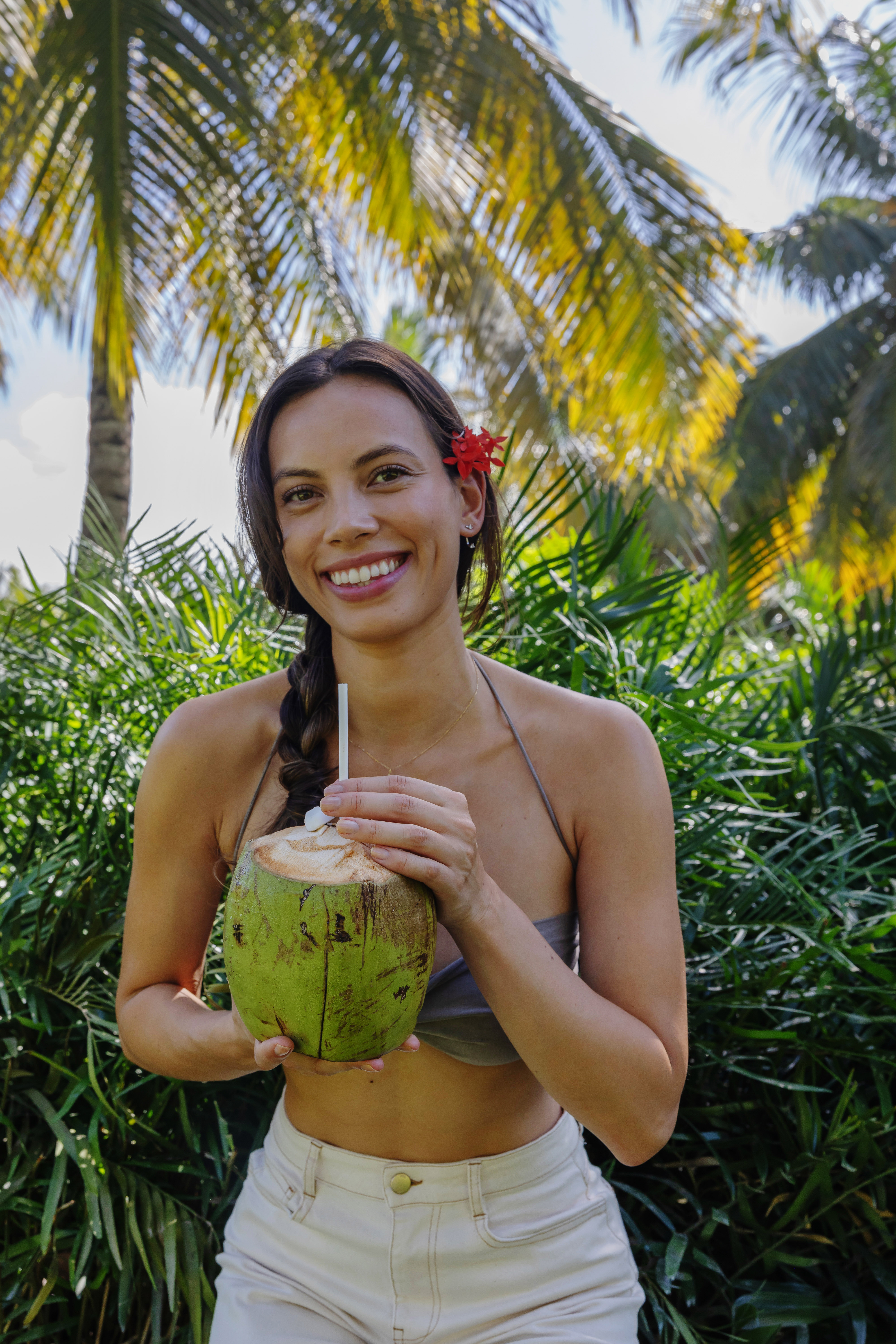 Mujer sonriendo, sosteniendo coco fresco con popote entre palmas tropicales.