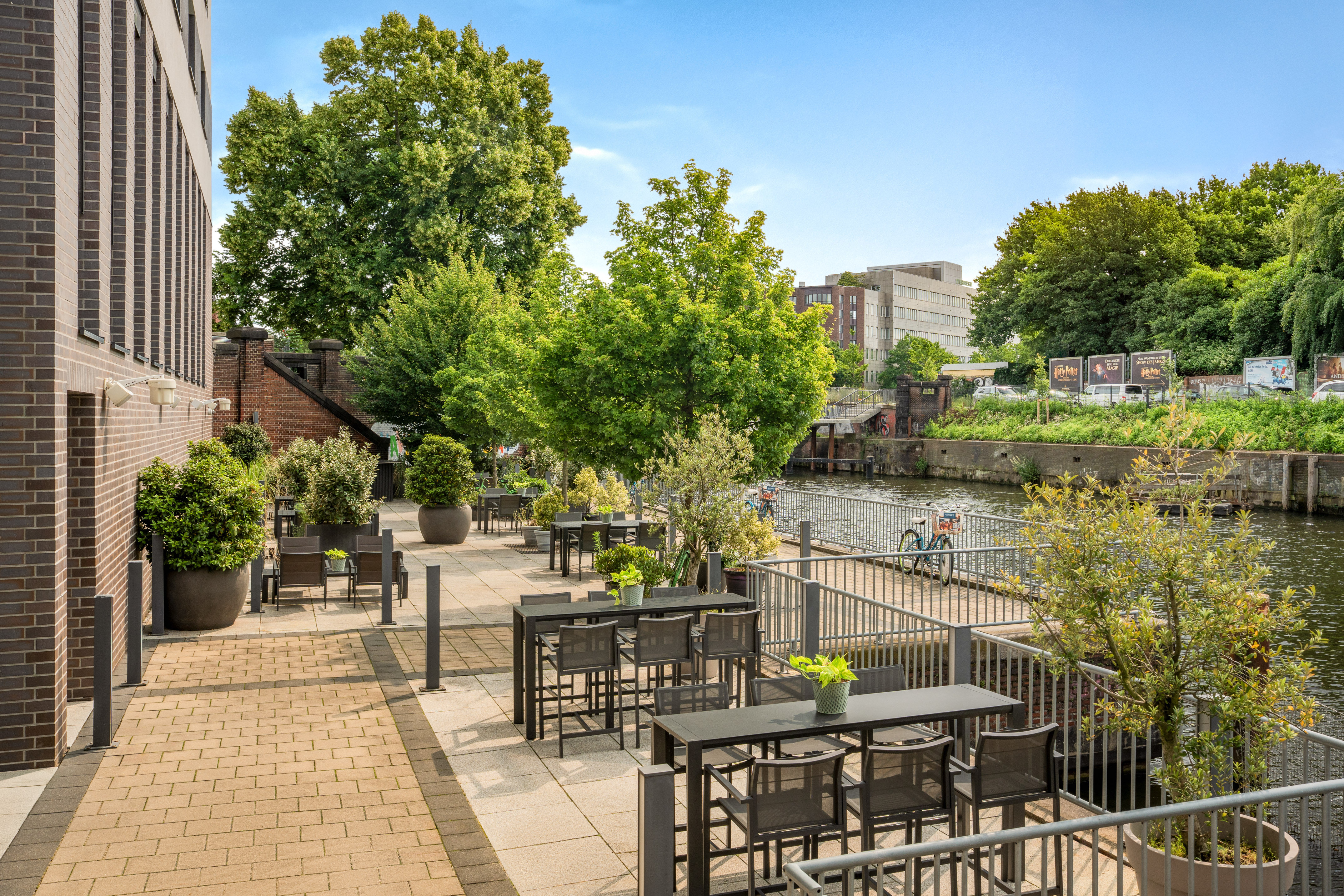 Modern outdoor patio by a canal with tables, chairs, and lush trees.