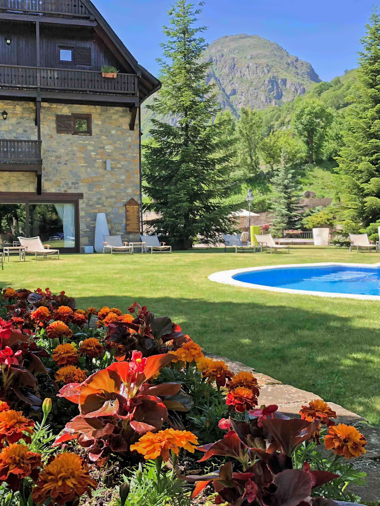 Hotel de montaña, piscina exterior, jardín con flores y vistas a la sierra.