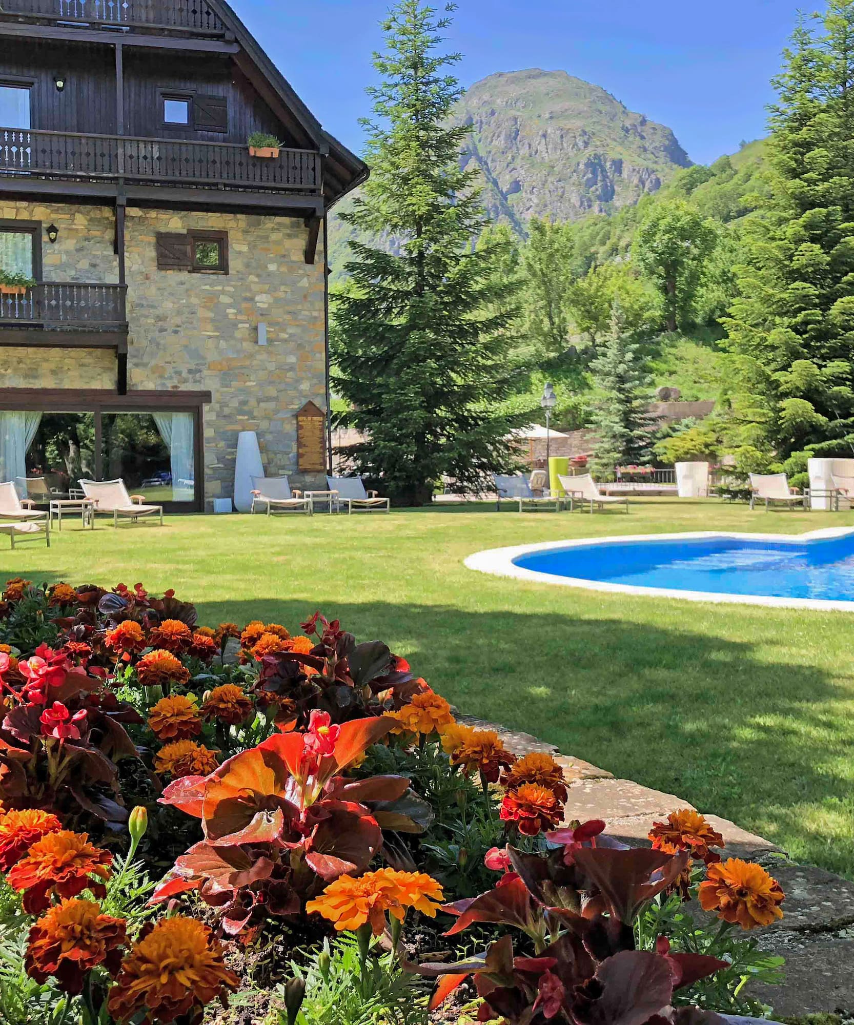 Hotel de montaña, piscina exterior, jardín con flores y vistas a la sierra.