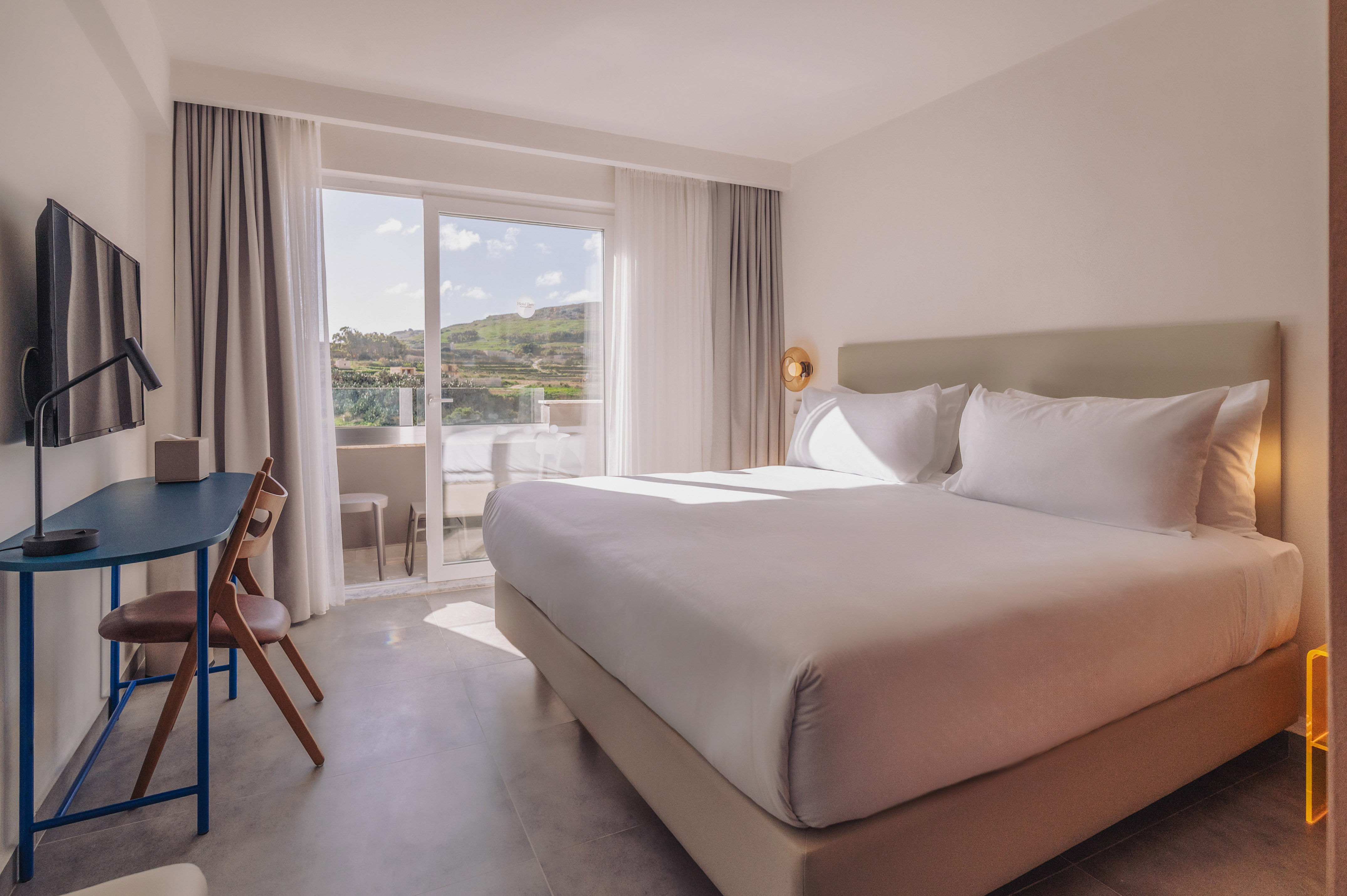 Modern hotel bedroom with bed, balcony, desk, and sunny hill view.