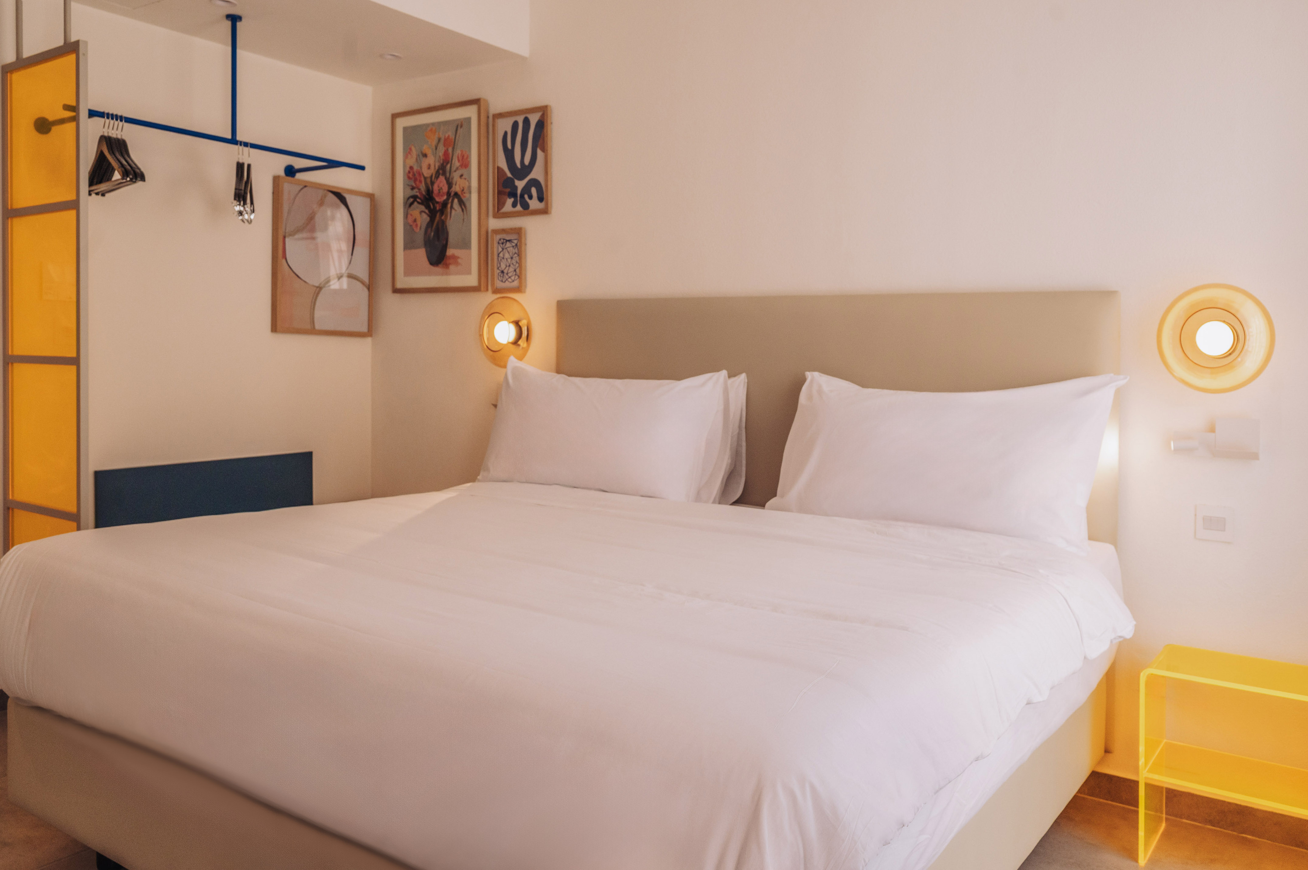 Modern minimalist hotel room with white bed, abstract art, and yellow accents.