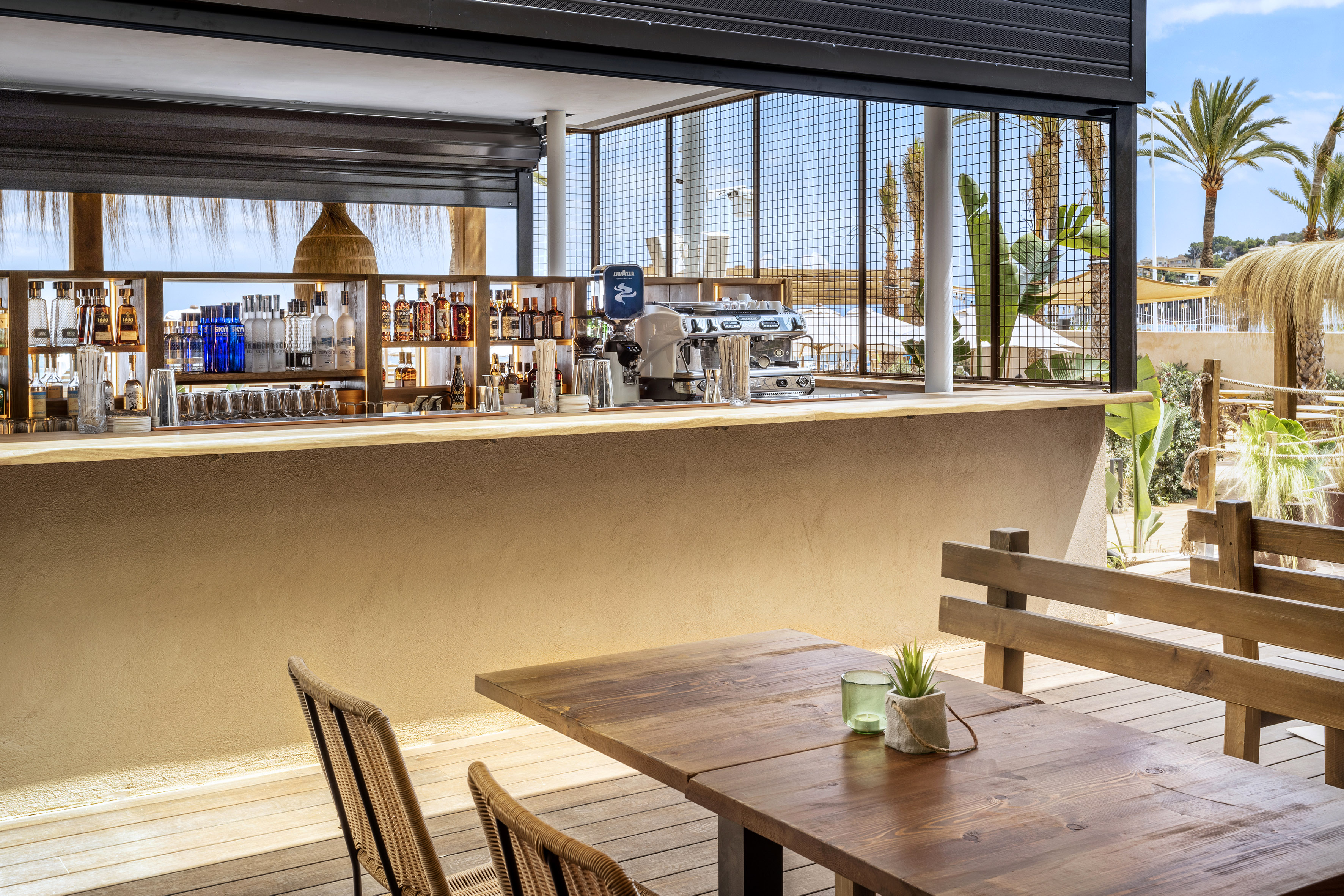 Resort outdoor bar with liquor bottles, coffee machine, wooden tables, palm trees.