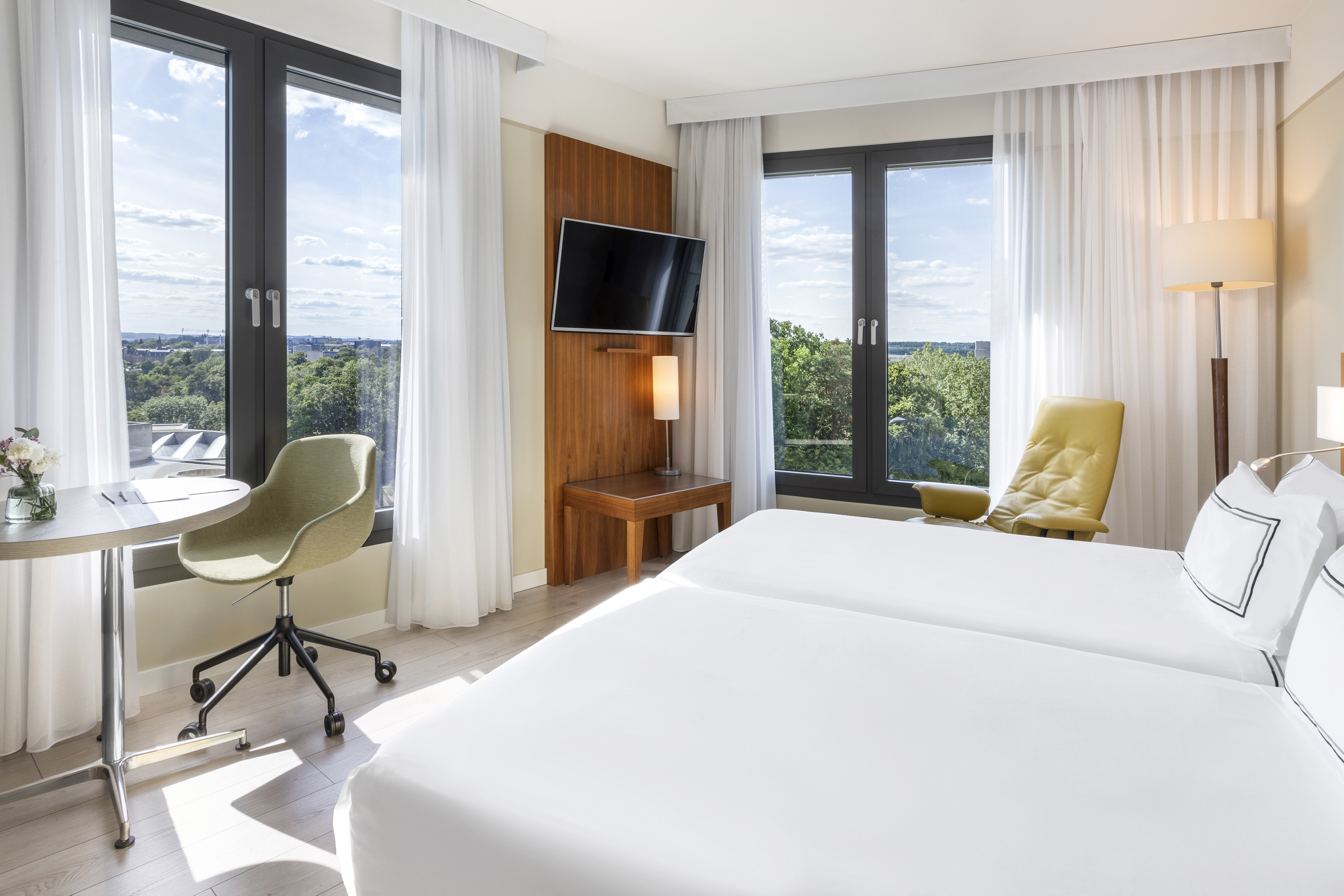 Bright hotel room with twin beds, city view windows, desk, and TV.