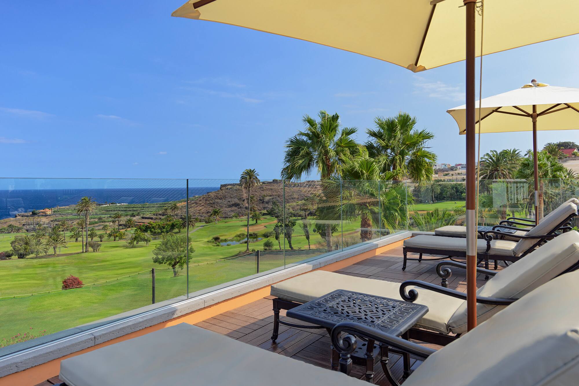 Terrace with lounge chairs, umbrellas, golf course, palm trees, and ocean view.
