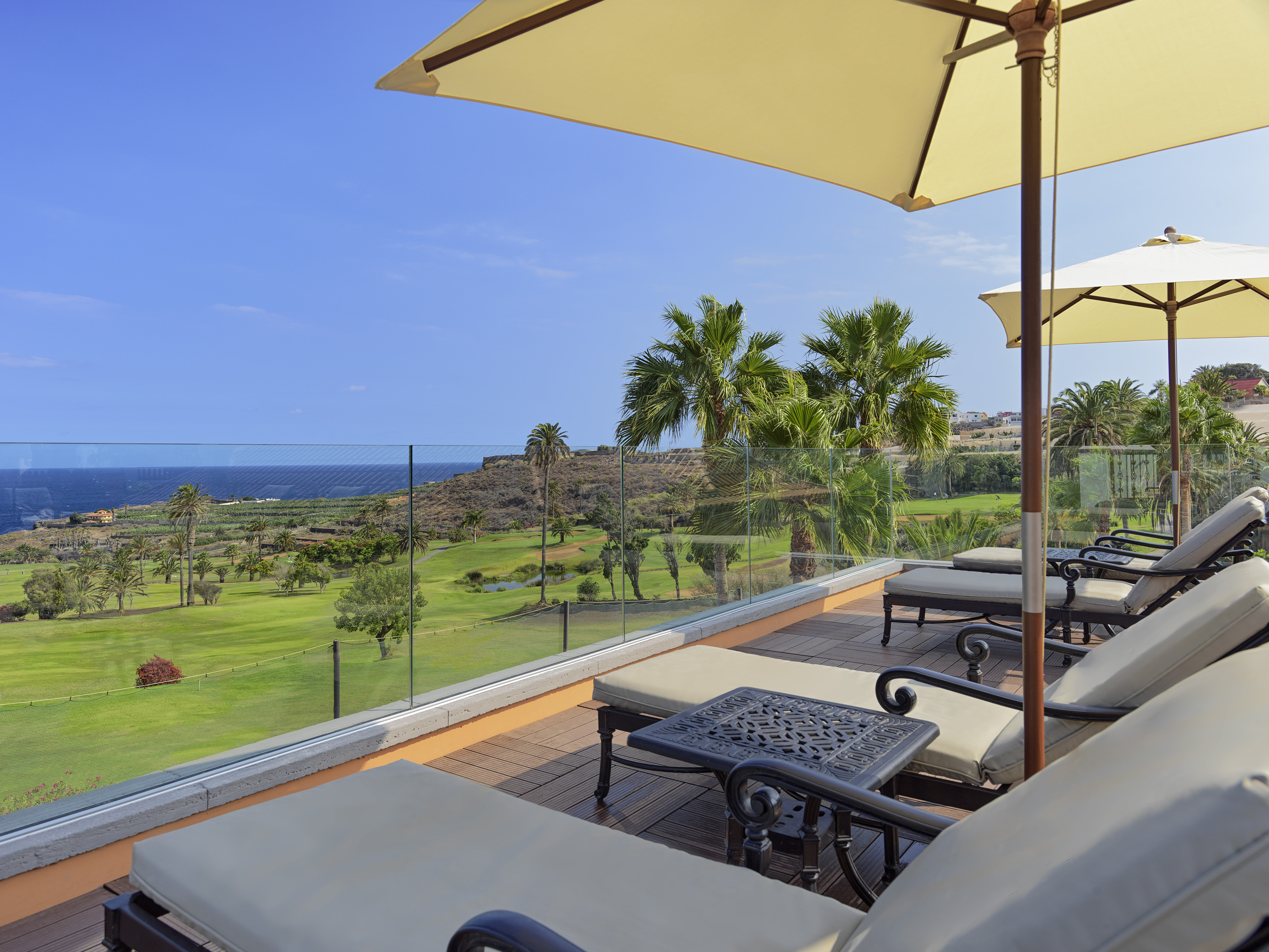 Terrace with lounge chairs, umbrellas, golf course, palm trees, and ocean view.