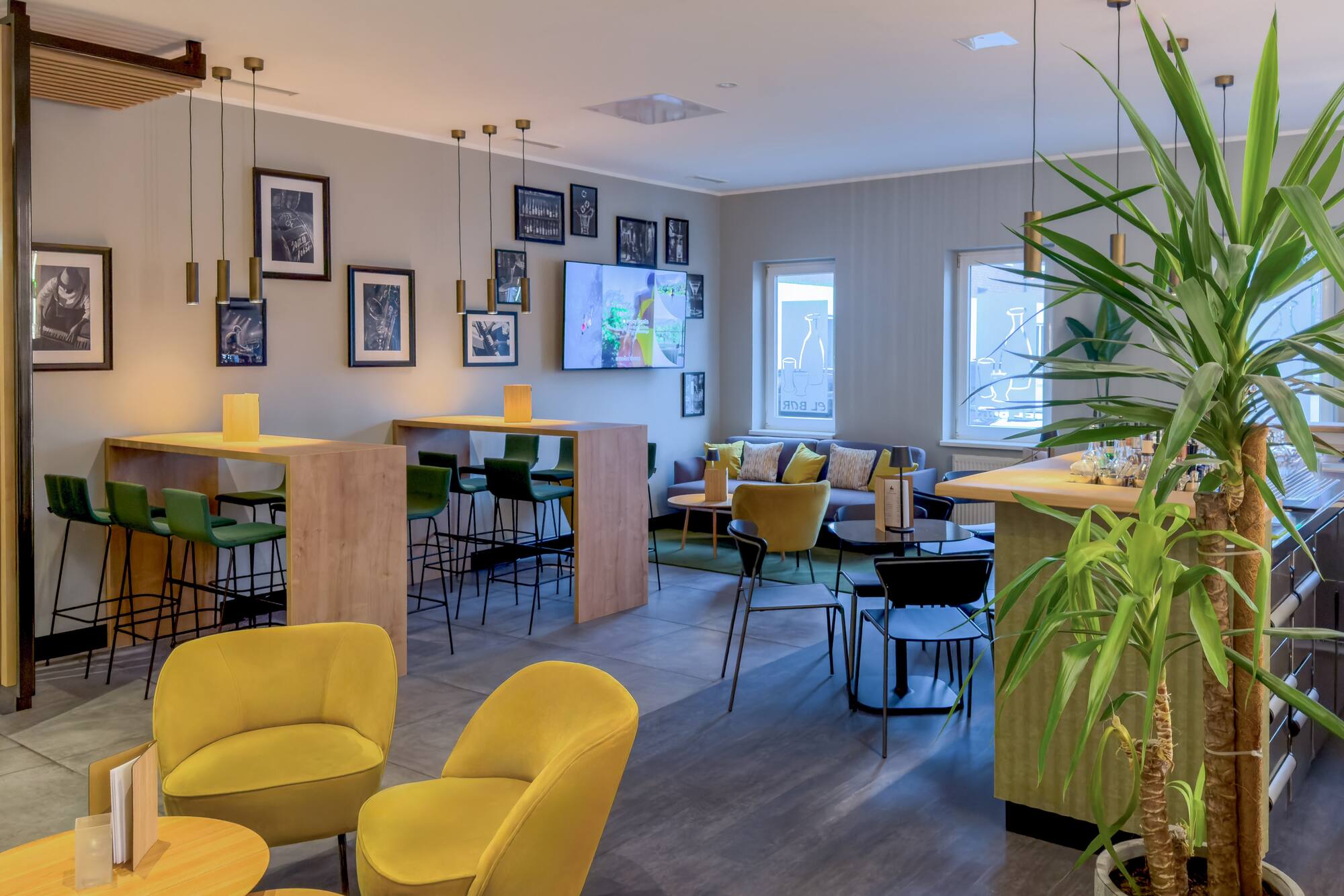 Modern hotel lounge bar with yellow chairs, green stools, framed art, and plants.