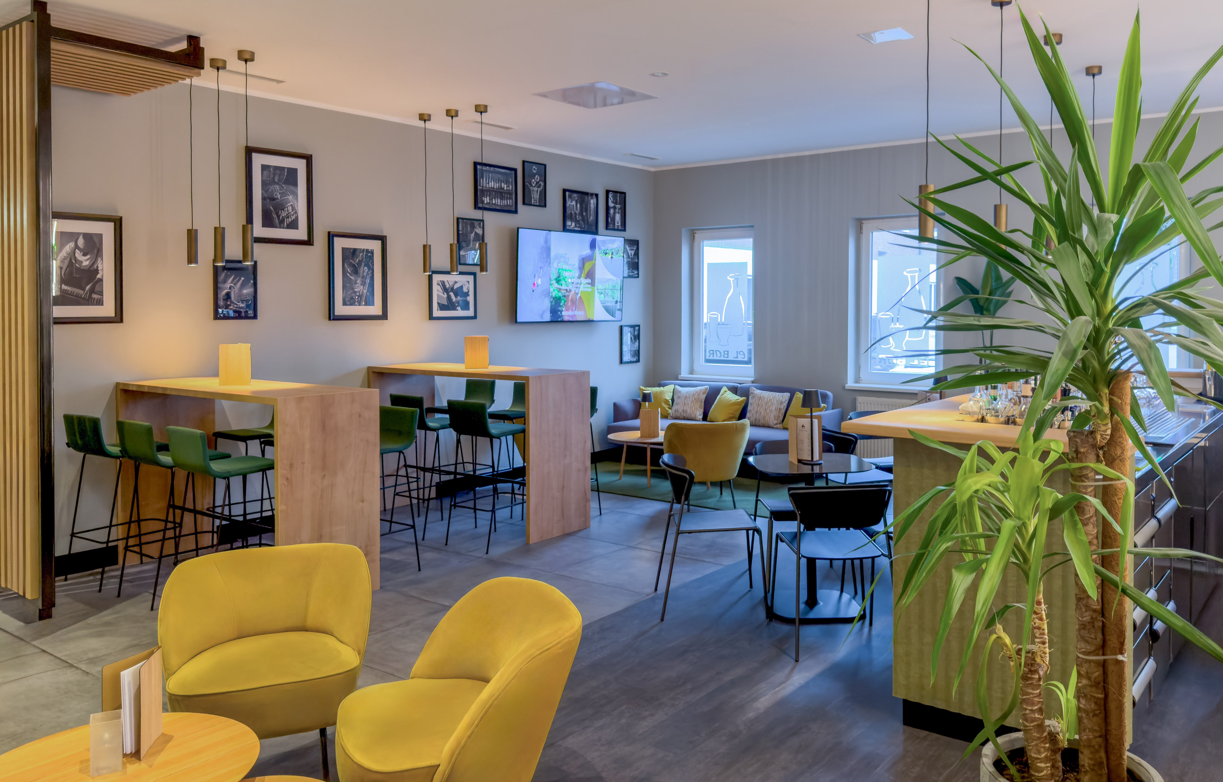 Modern hotel lounge bar with yellow chairs, green stools, framed art, and plants.