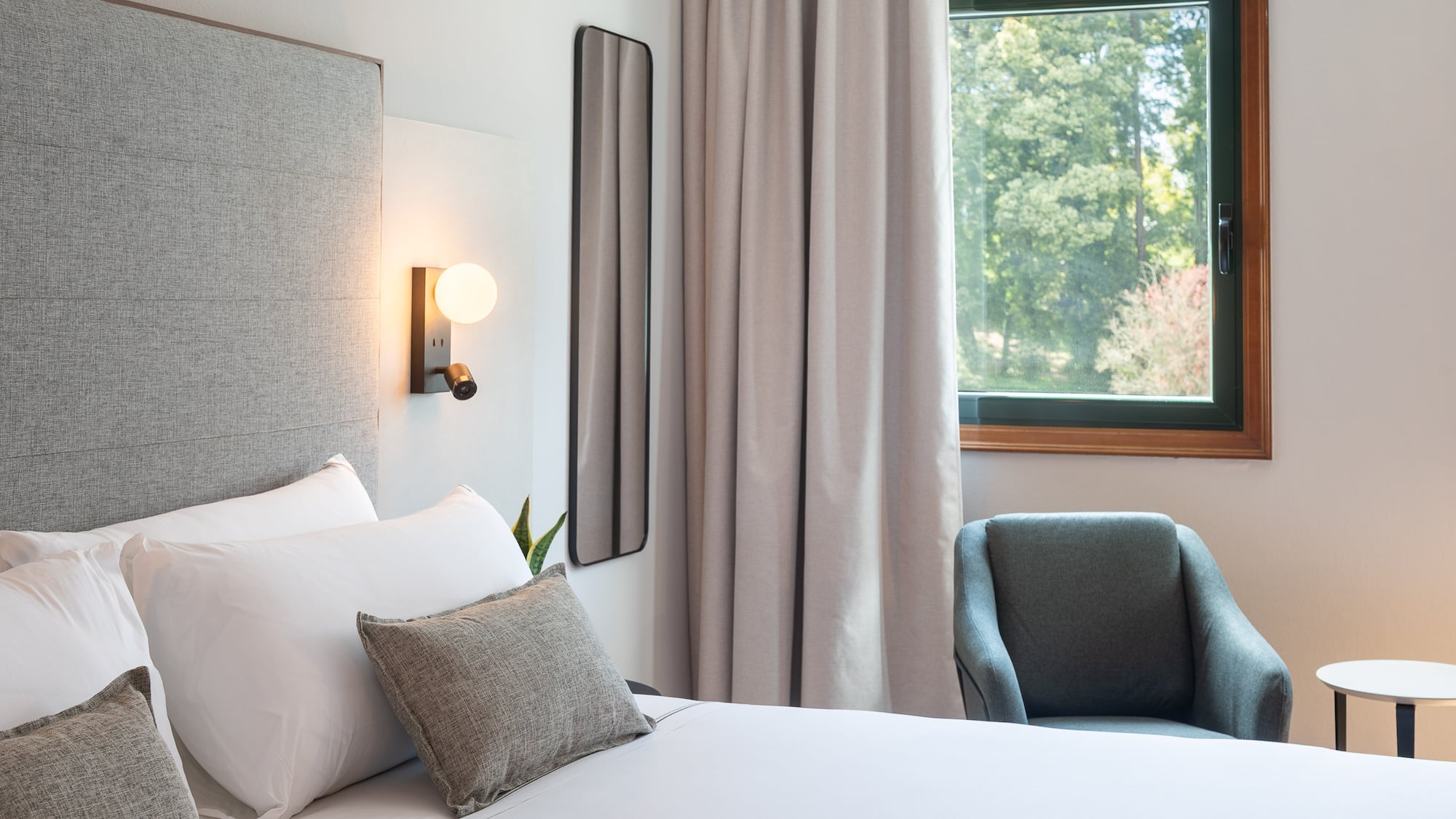 Modern hotel room with comfy bed, grey armchair, and window overlooking trees.