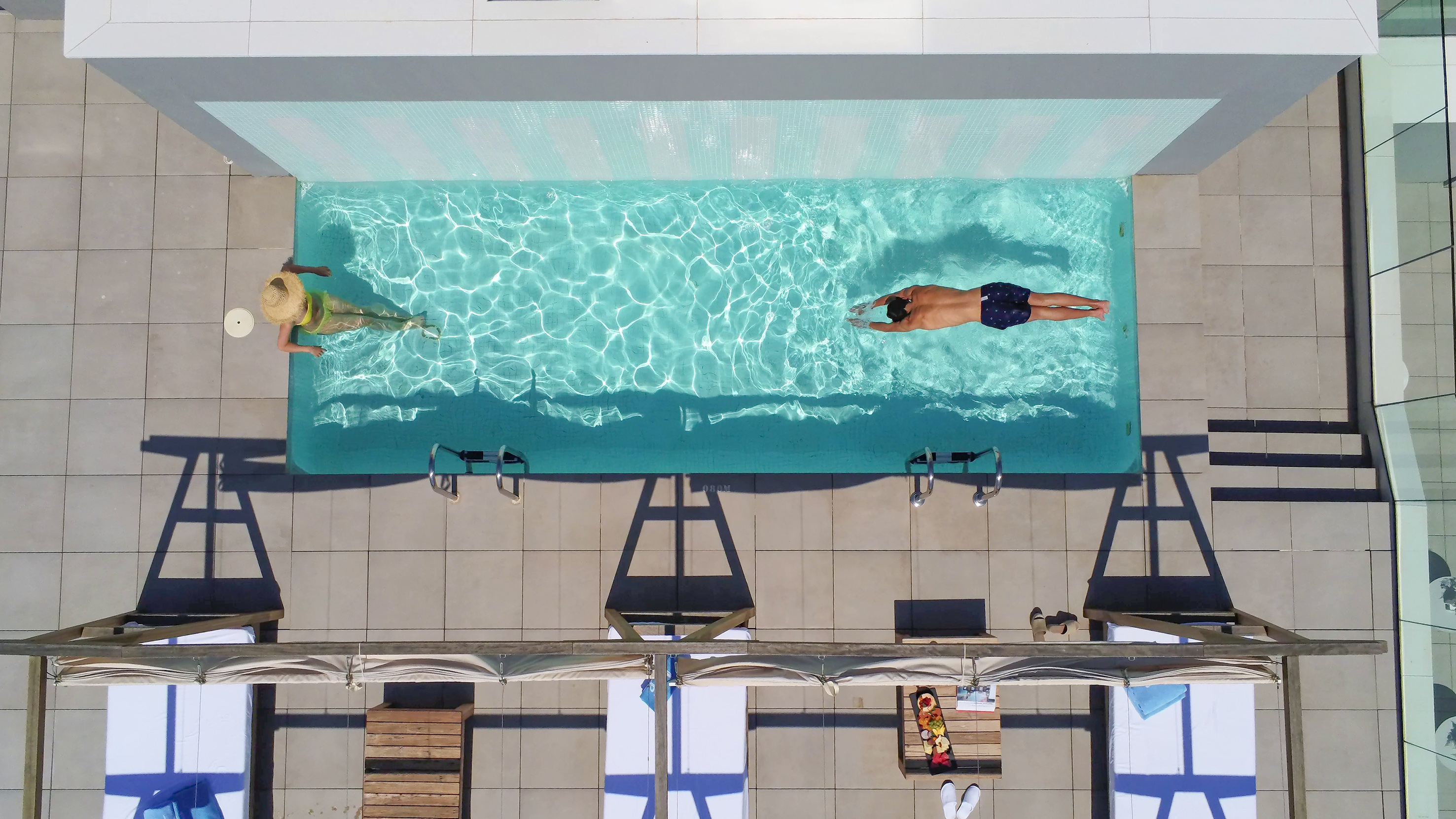 Aerial view of two people swimming in a resort pool with patio lounge chairs.