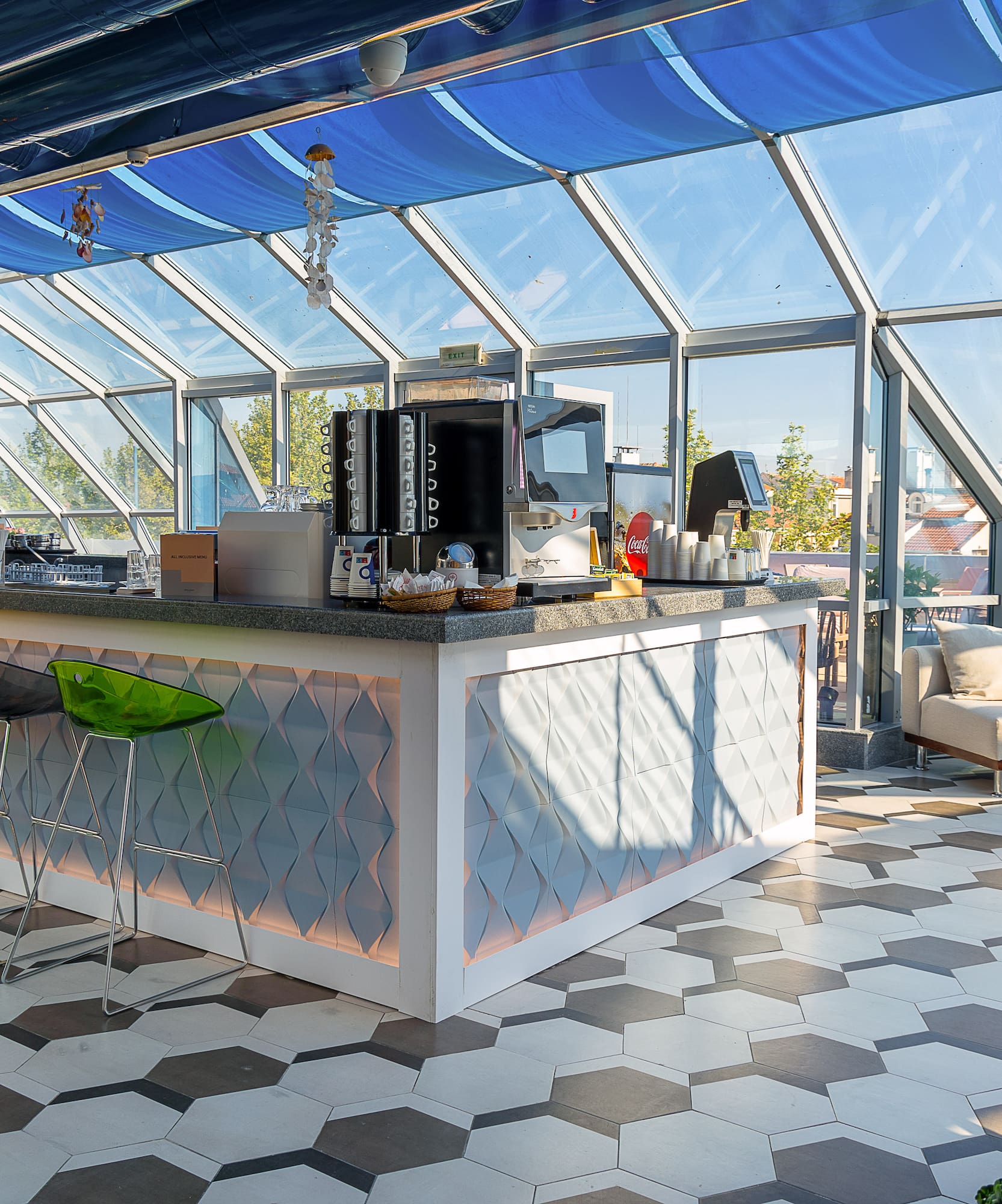 Modern rooftop bar with glass walls, blue canopy, colorful stools, and lounge seating.