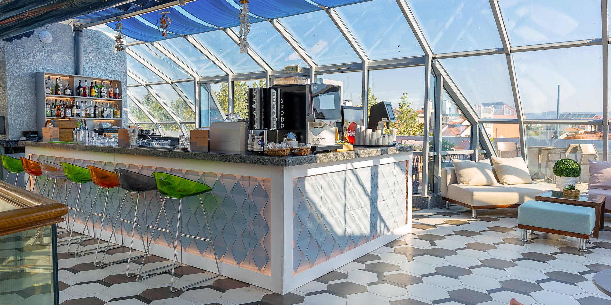 Modern rooftop bar with glass walls, blue canopy, colorful stools, and lounge seating.