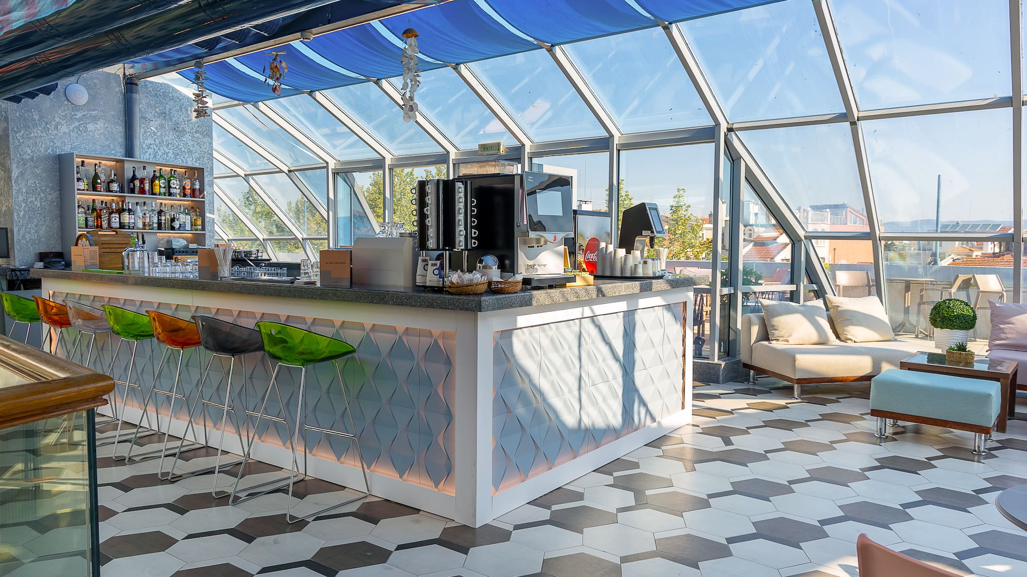 Modern rooftop bar and lounge with colorful stools, glass roof, and hexagon floor.