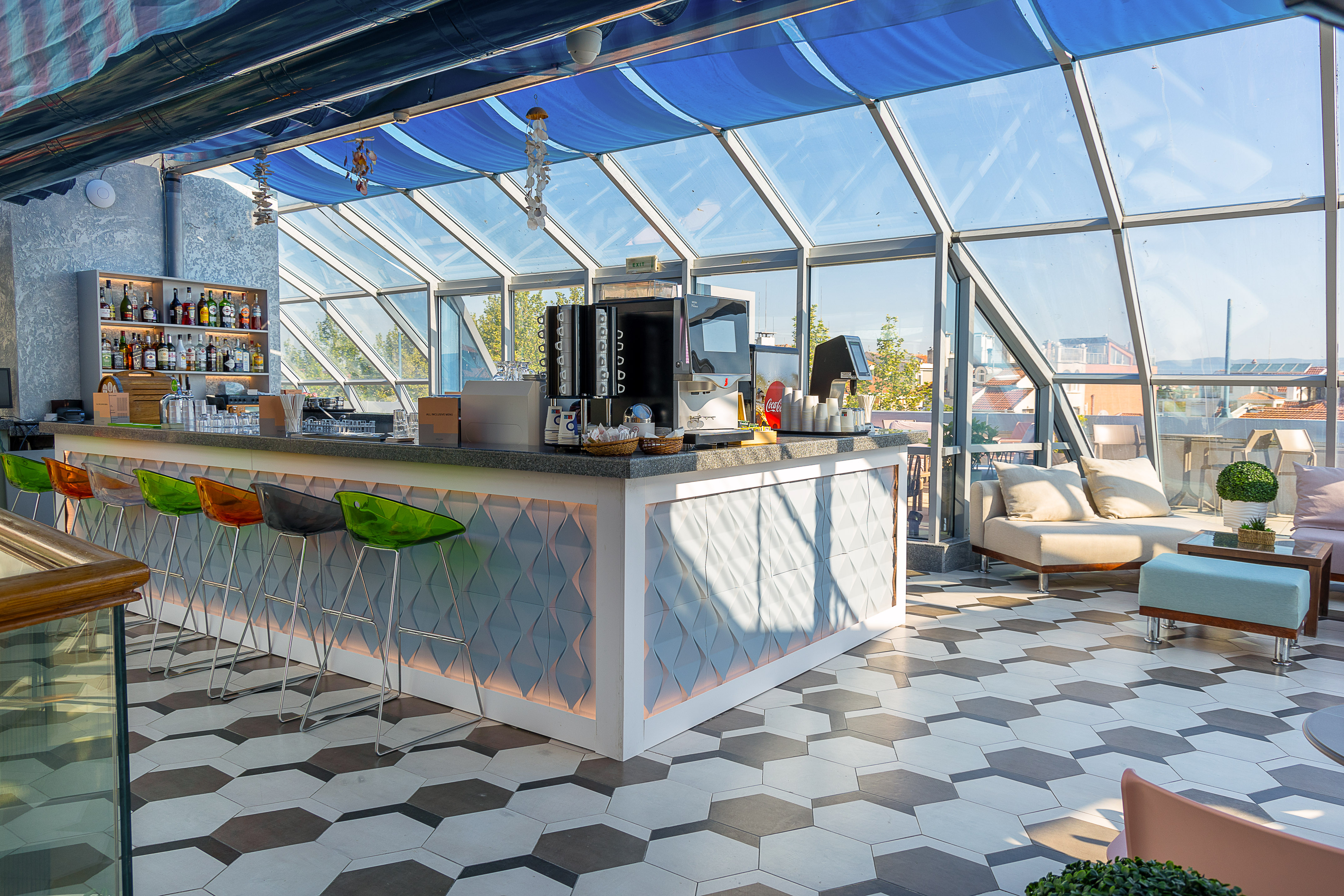 Modern rooftop bar with glass walls, blue canopy, colorful stools, and lounge seating.