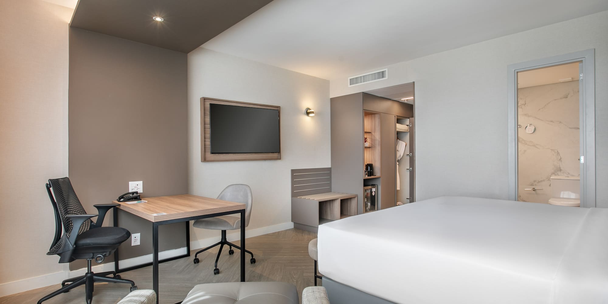 Modern hotel guest room with bed, work desk, wall TV, and open wardrobe.
