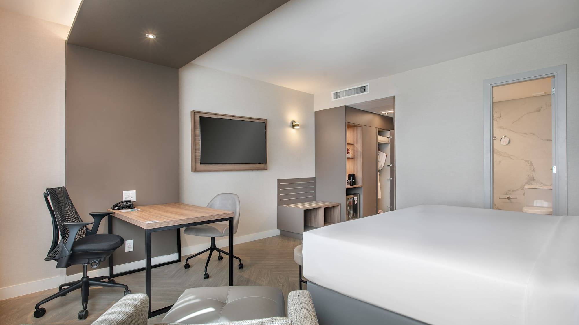 Contemporary hotel room with king bed, work desk, wall TV, and open closet.