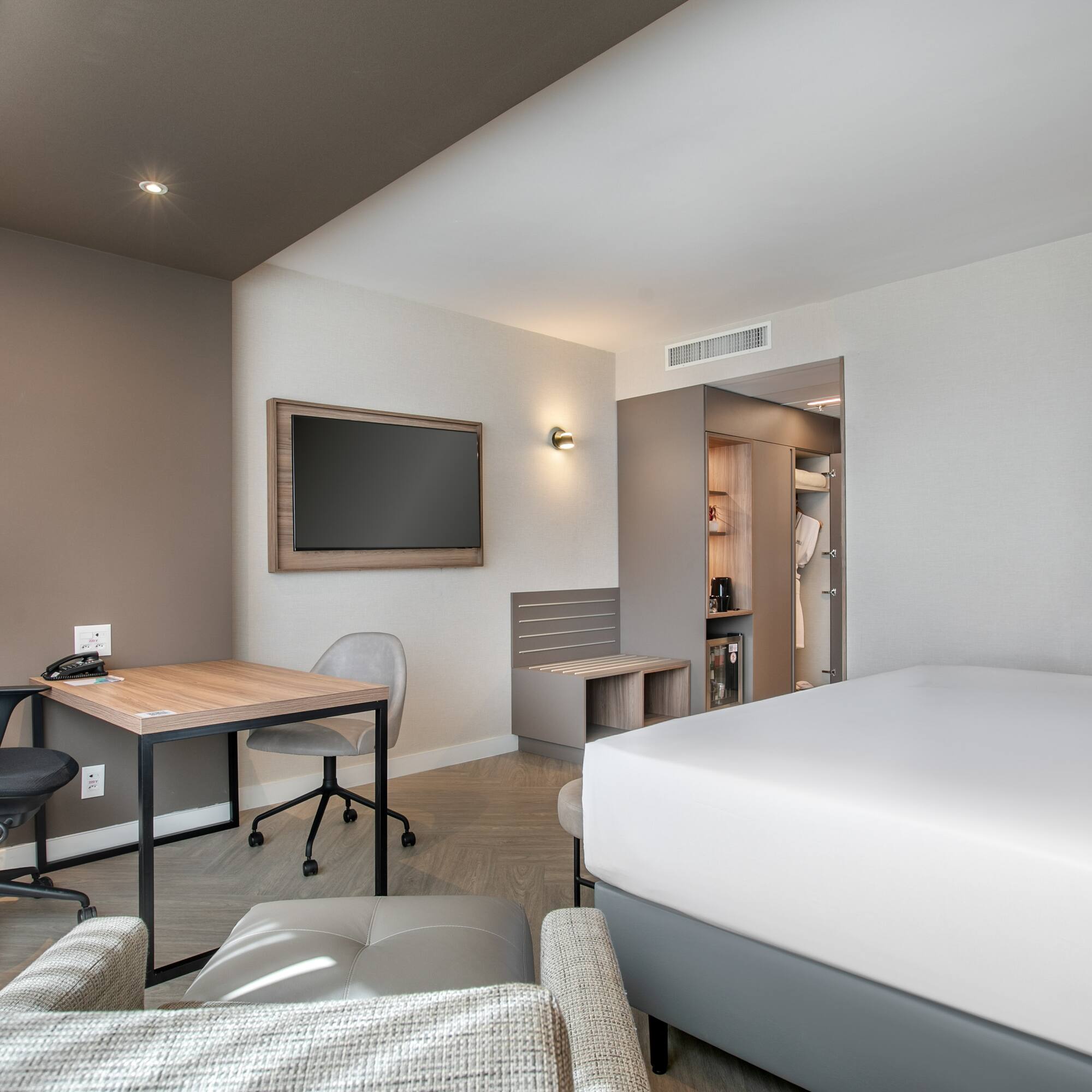 Modern hotel guest room with bed, work desk, wall TV, and open wardrobe.