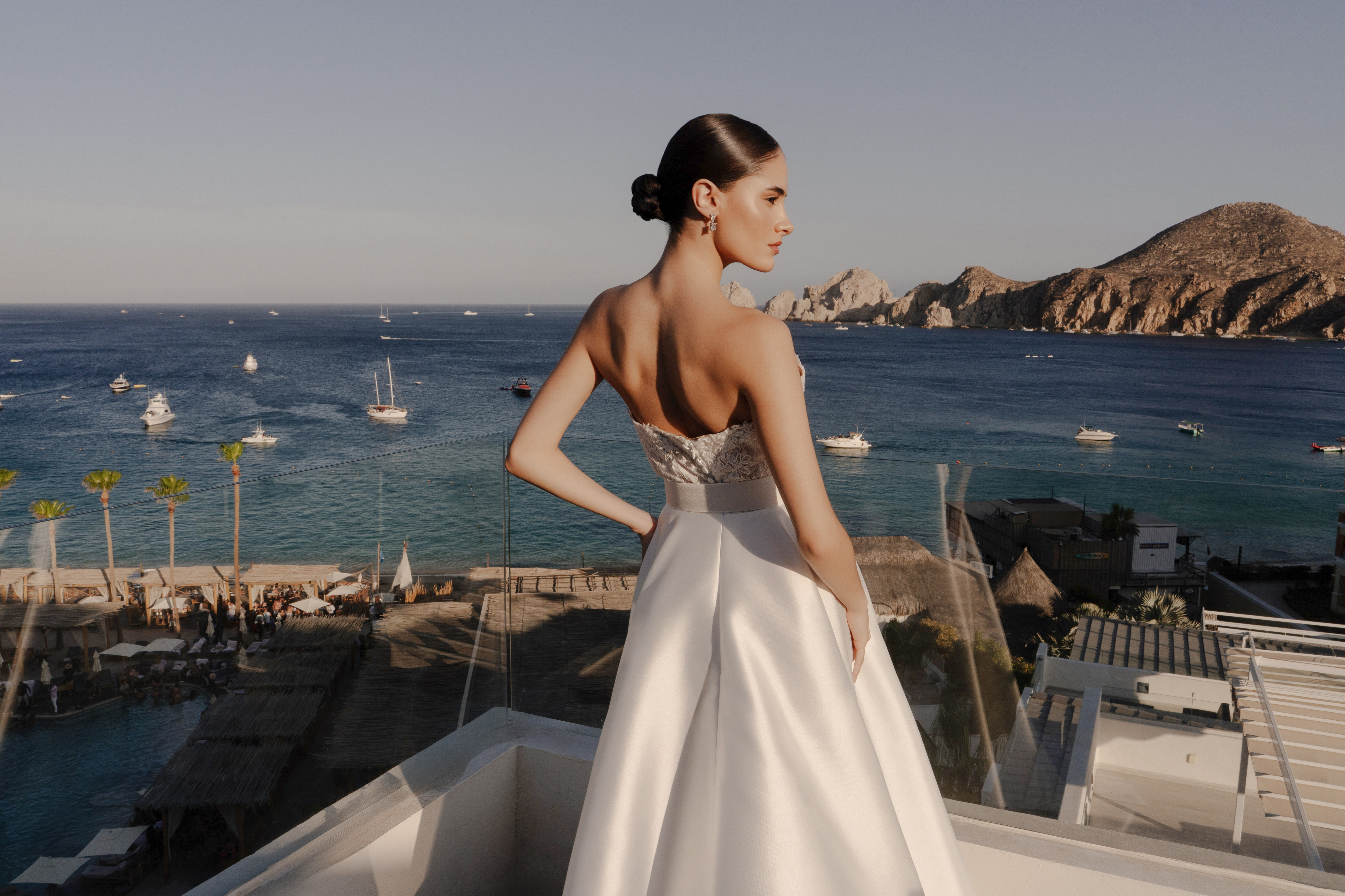 Woman in white bridal gown on balcony, ocean view with boats and rocky coast.