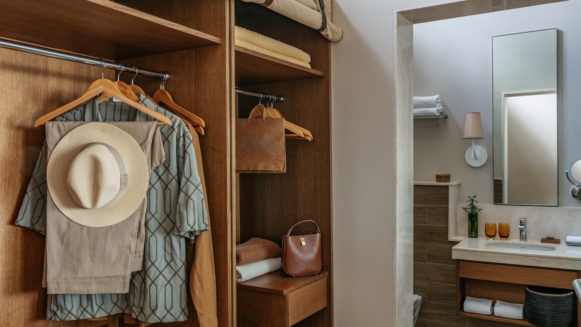 Wooden hotel closet with hanging clothes, hat, towels, and modern bathroom.