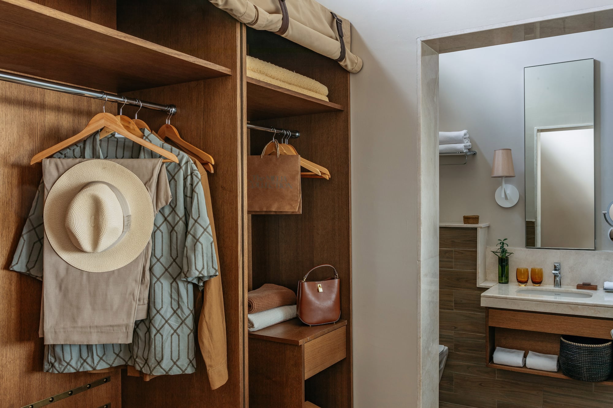Wooden hotel closet with hanging clothes, hat, towels, and modern bathroom.