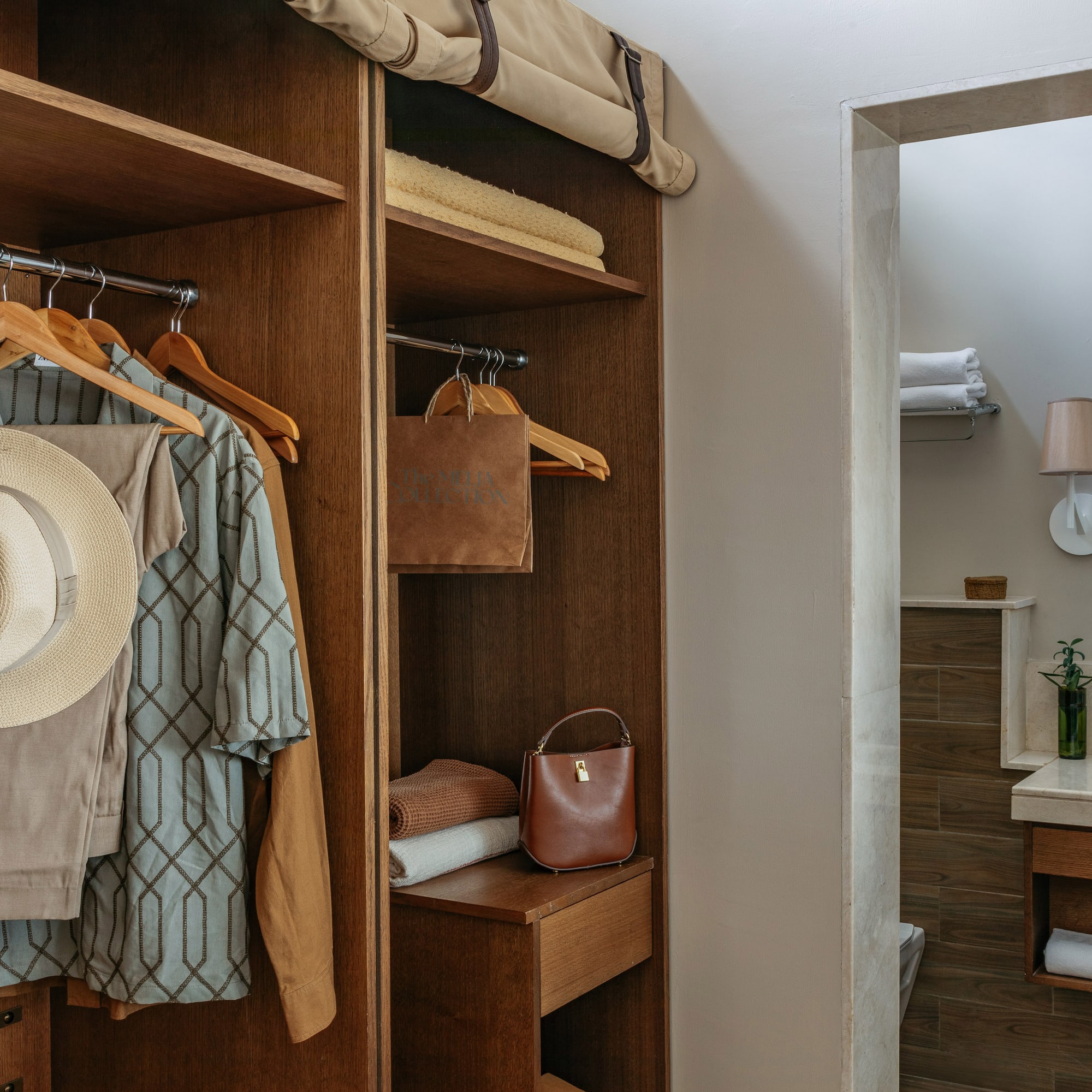 Wooden hotel closet with hanging clothes, hat, towels, and modern bathroom.