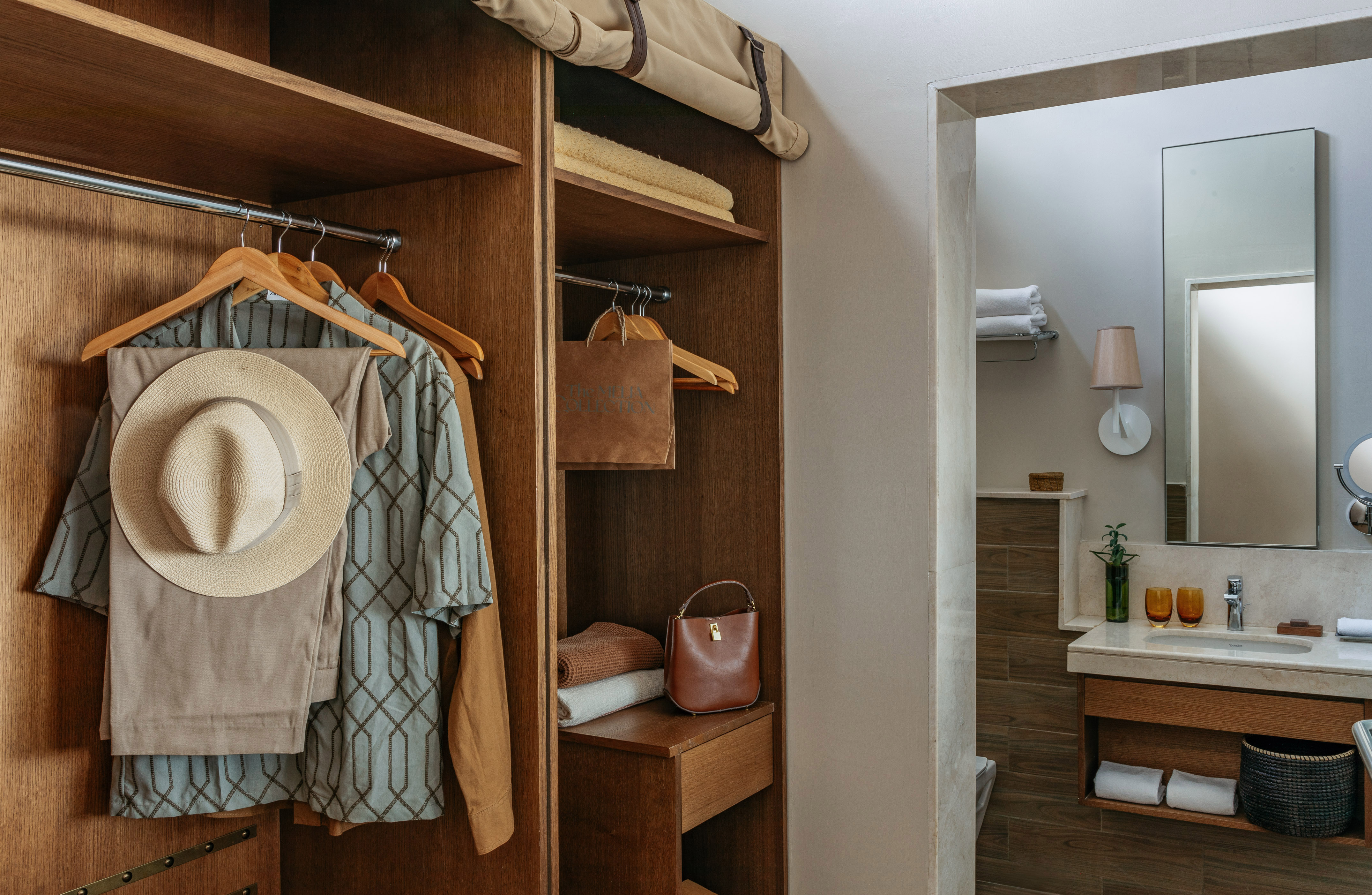 Wooden hotel closet with hanging clothes, hat, towels, and modern bathroom.
