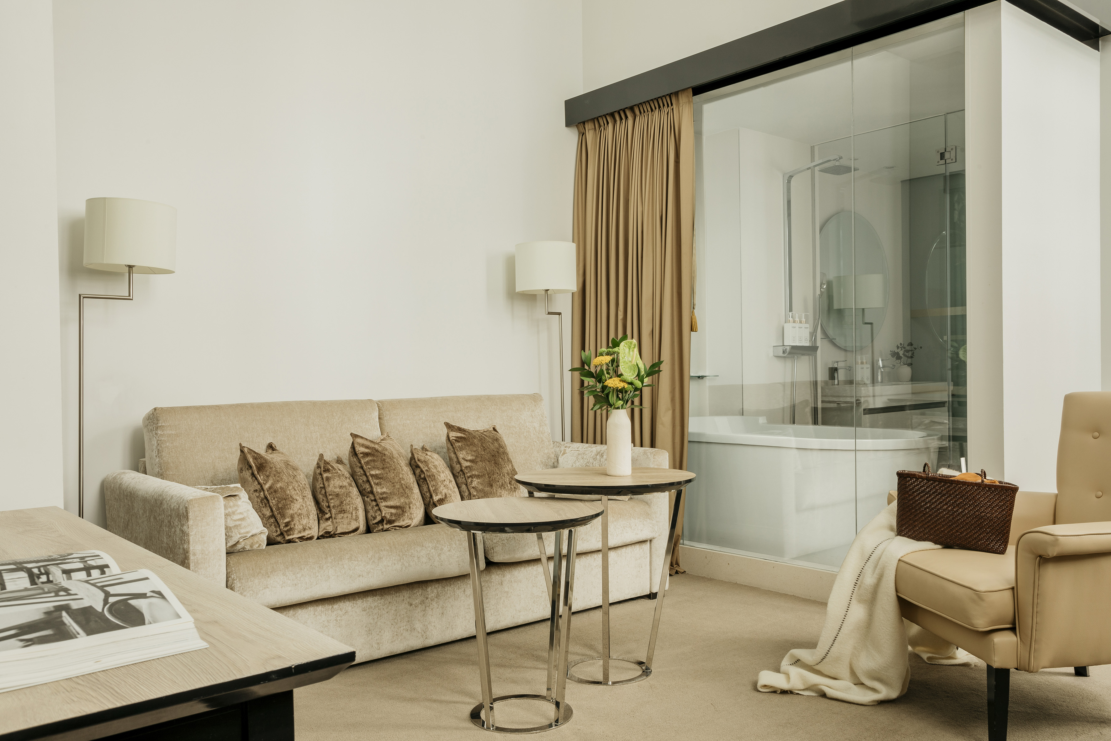 Modern hotel suite living area with beige couch and glass bathroom.