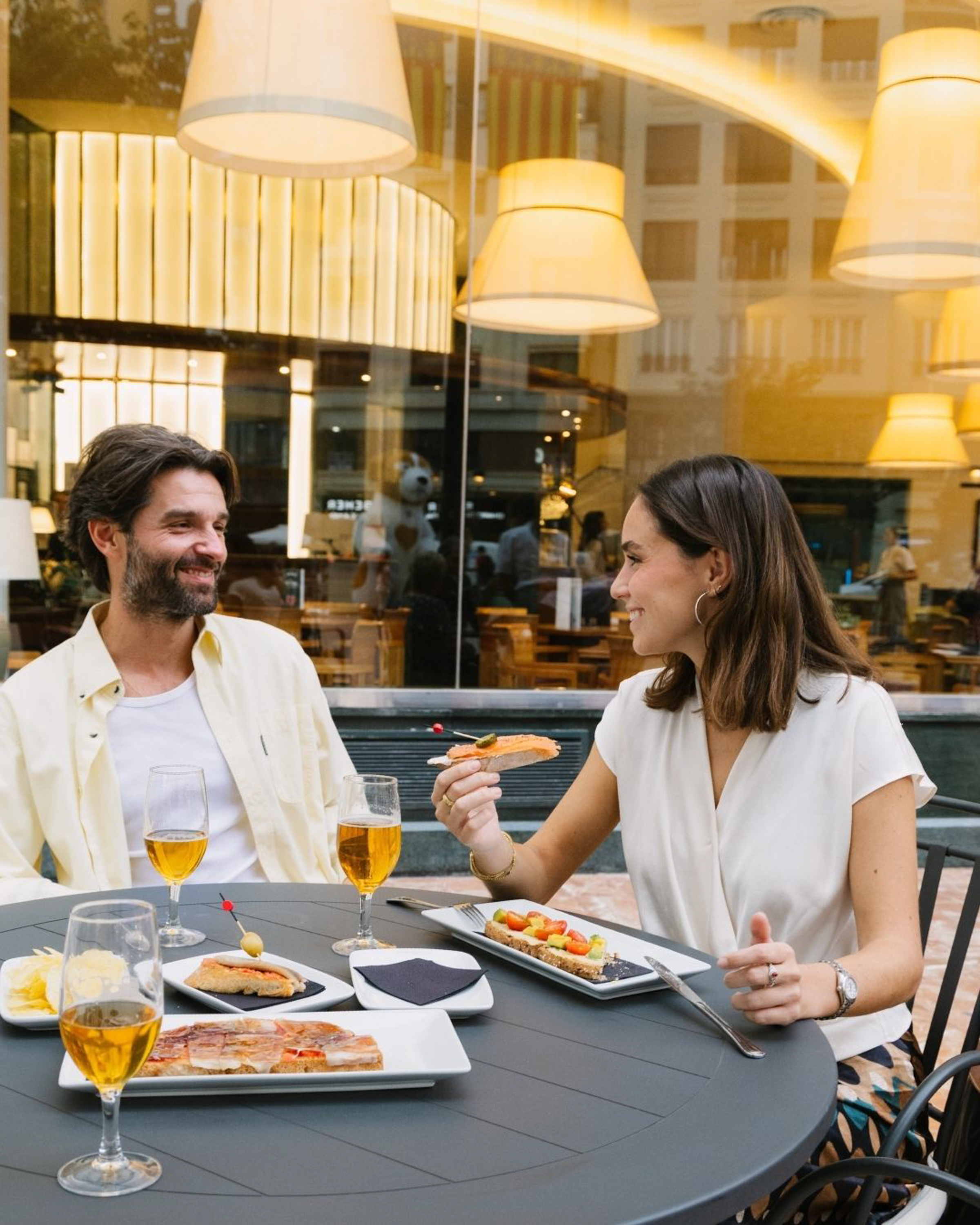 Couple smiling, enjoying tapas and beer at an outdoor restaurant.