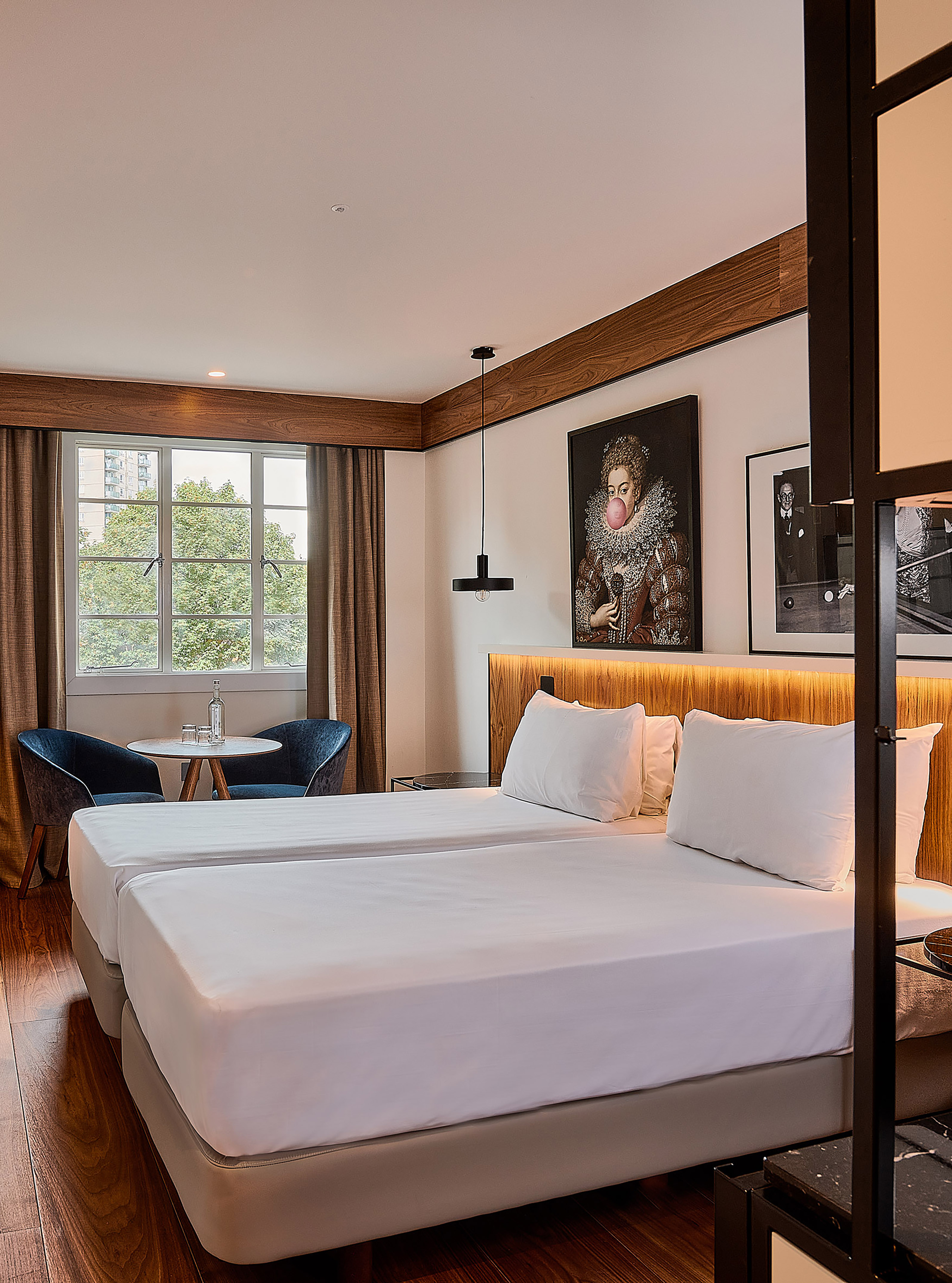 Hotel room with twin beds, wooden floors, city view, and unique wall art.