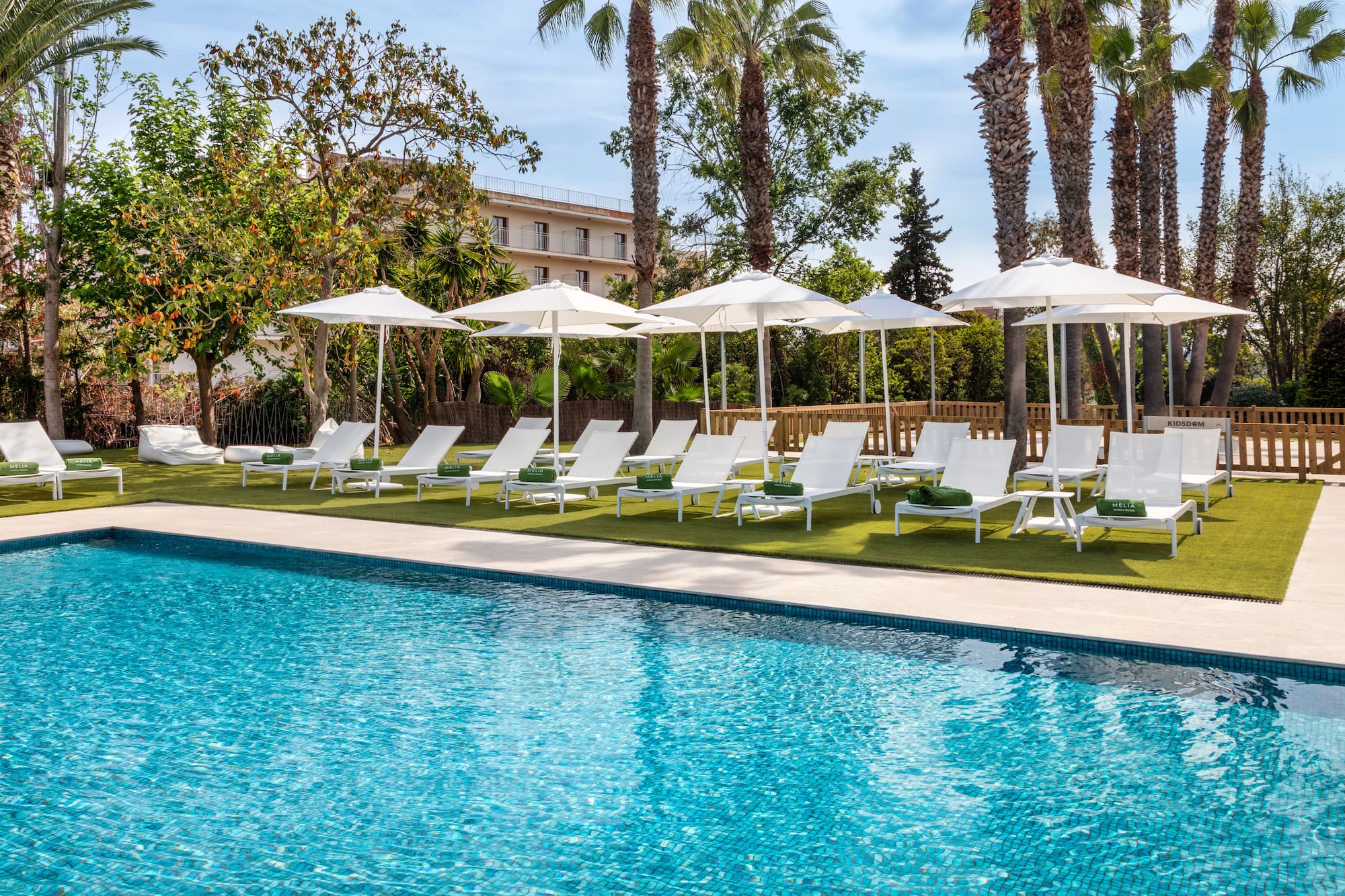 Resort swimming pool with white lounge chairs, umbrellas, and palm trees.