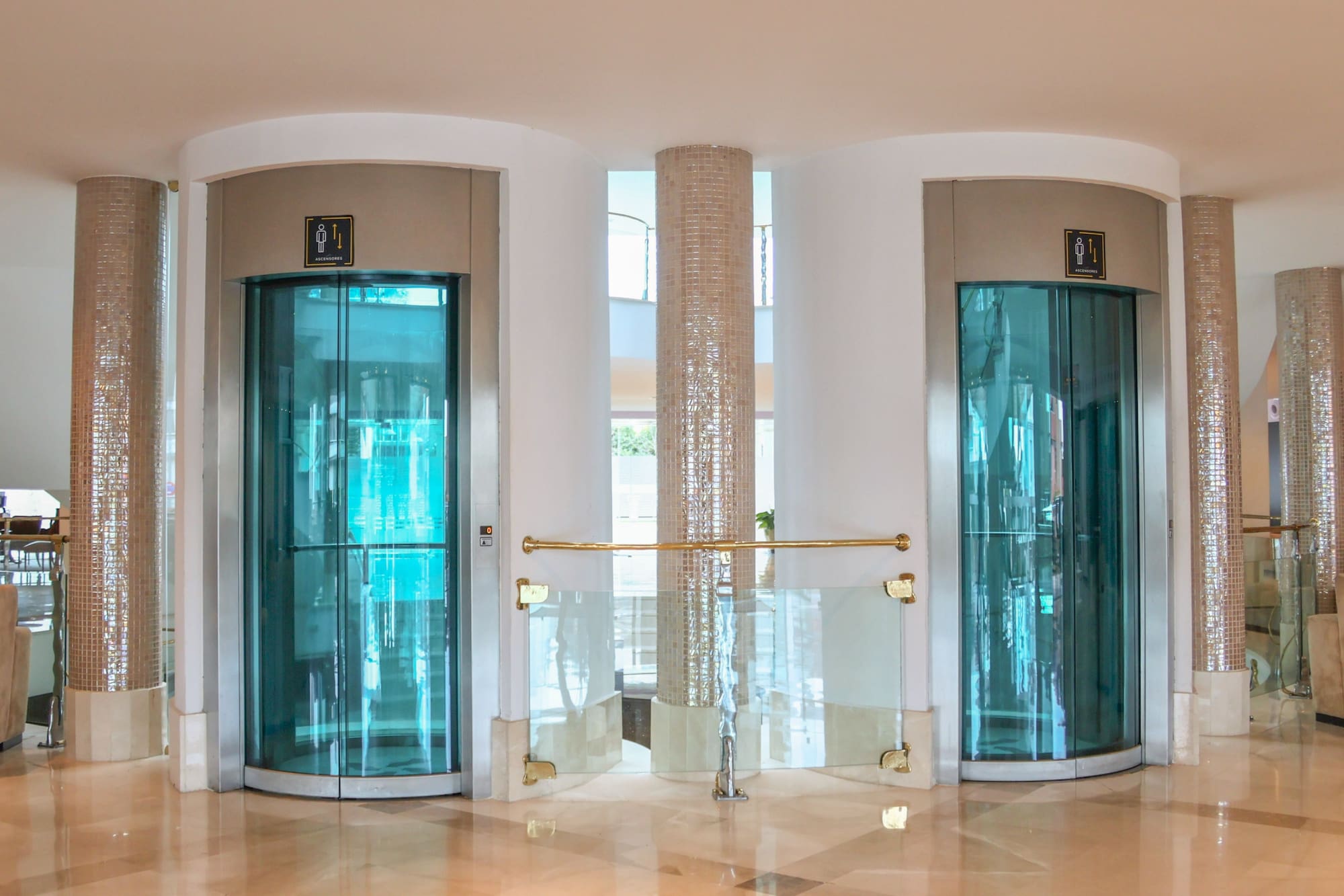 Two modern glass elevators in a luxury hotel lobby with tiled pillars.