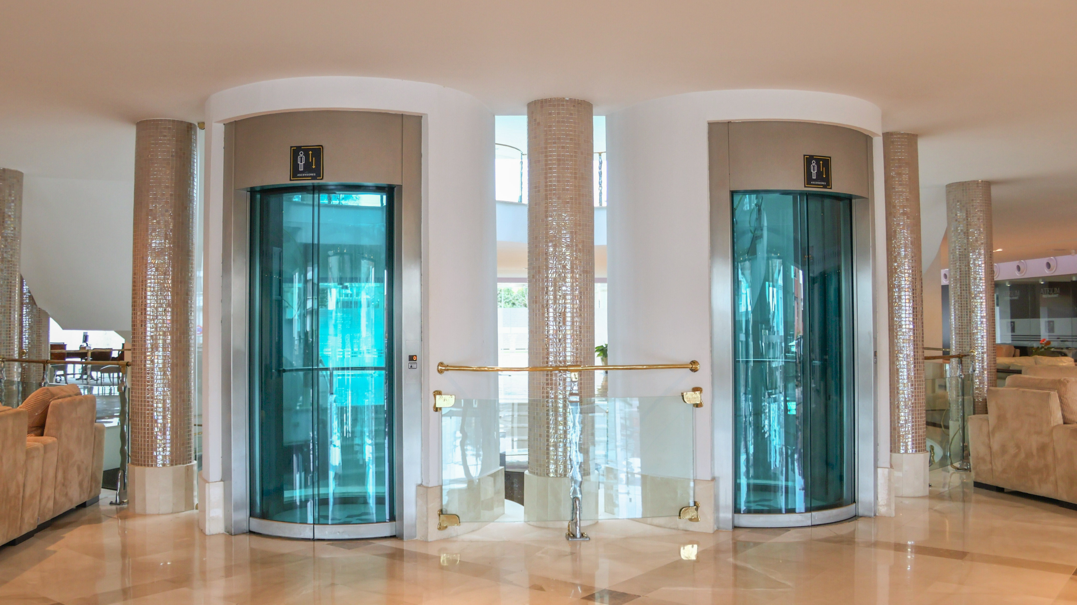 Two modern glass elevators in a luxury hotel lobby with tiled pillars.