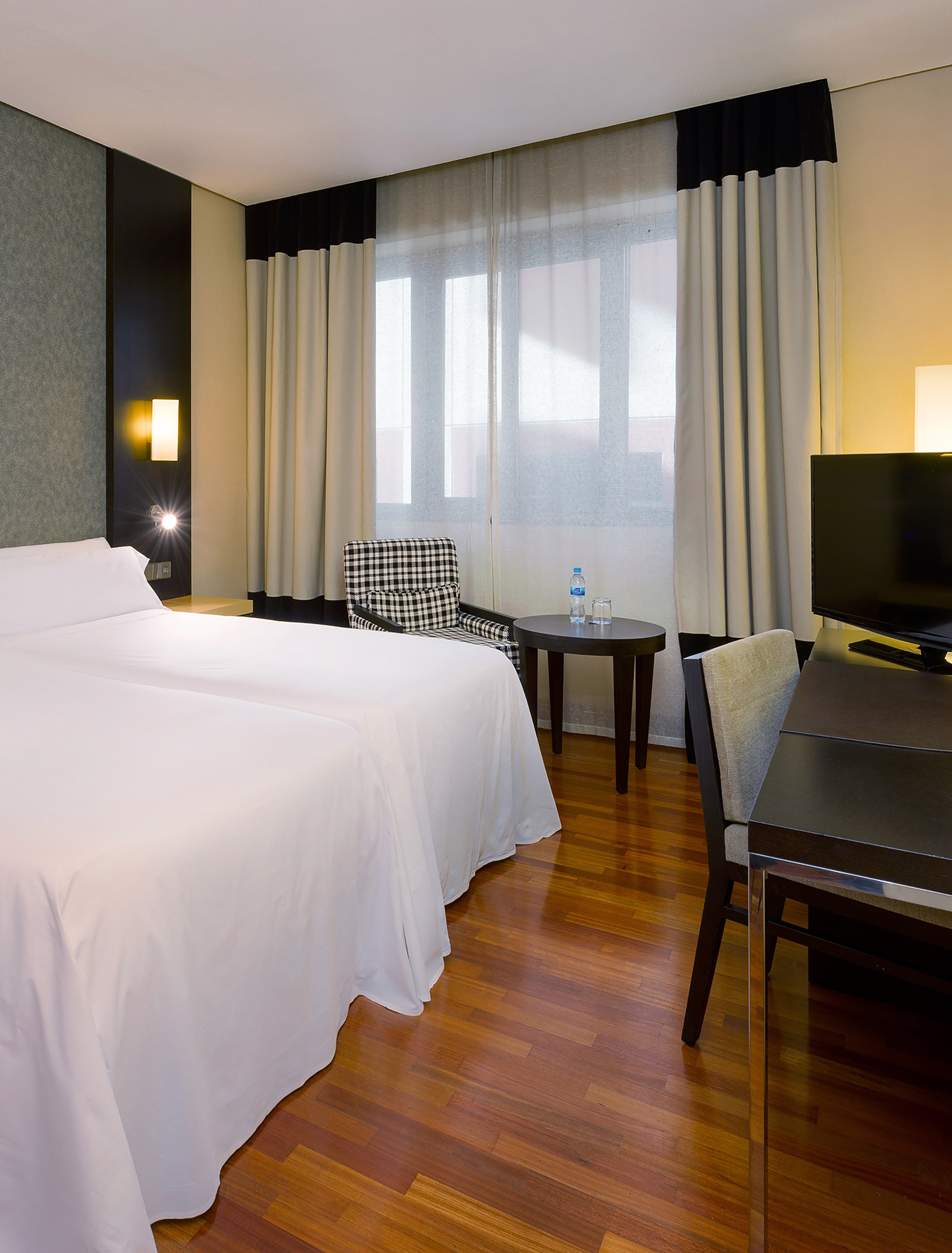 Modern hotel room with twin beds, wooden floor, desk, TV, and checkered armchair.