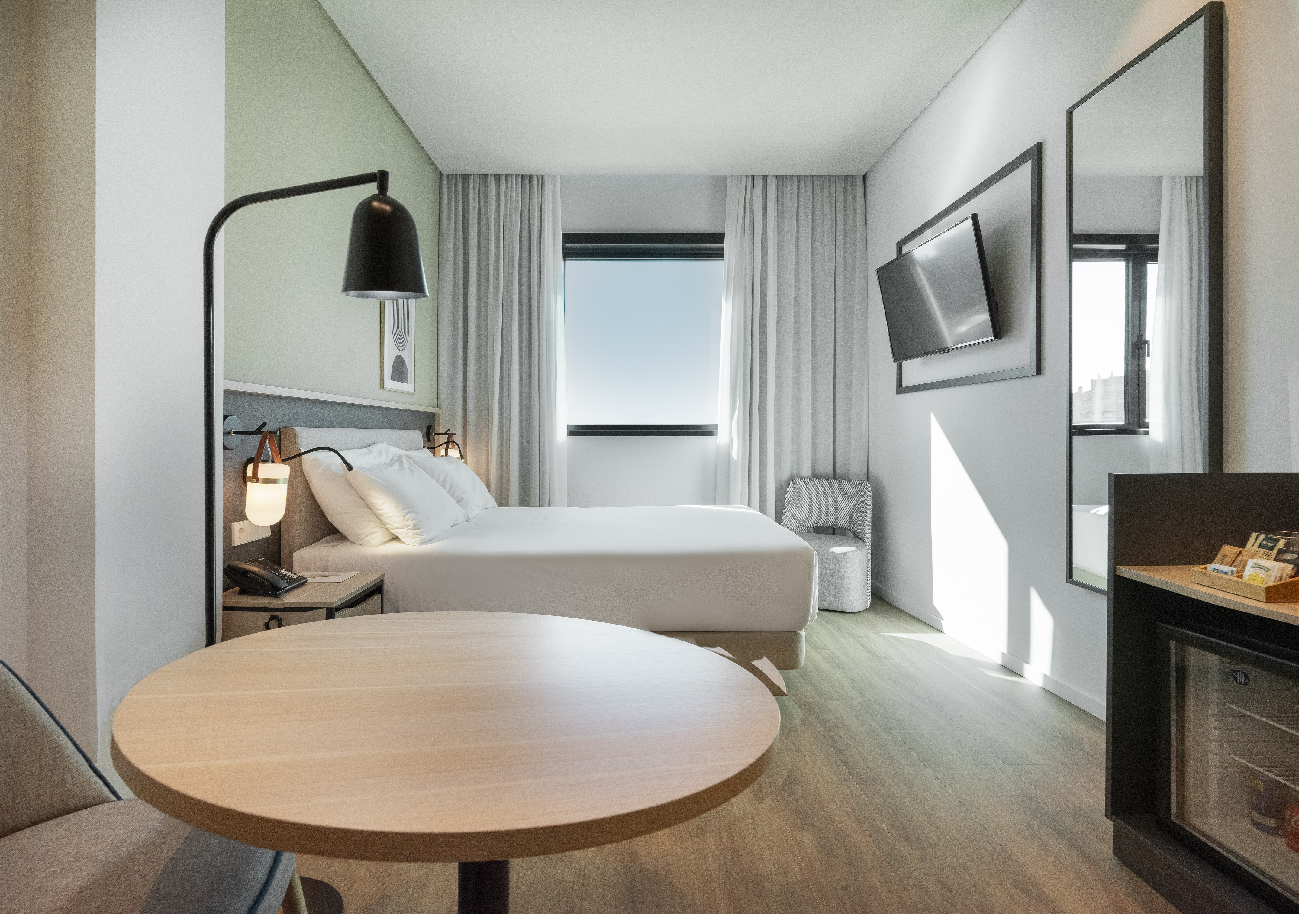 Modern hotel room with white bed, round table, wall TV, and minibar.
