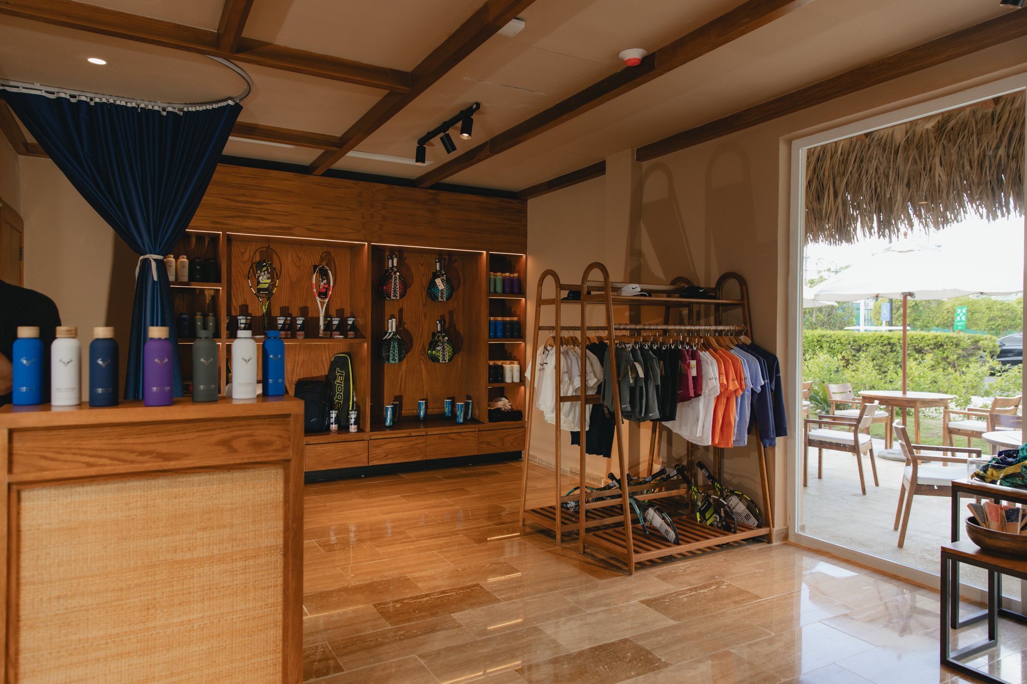 Modern pro shop with clothing, tennis rackets, water bottles, and outdoor view.