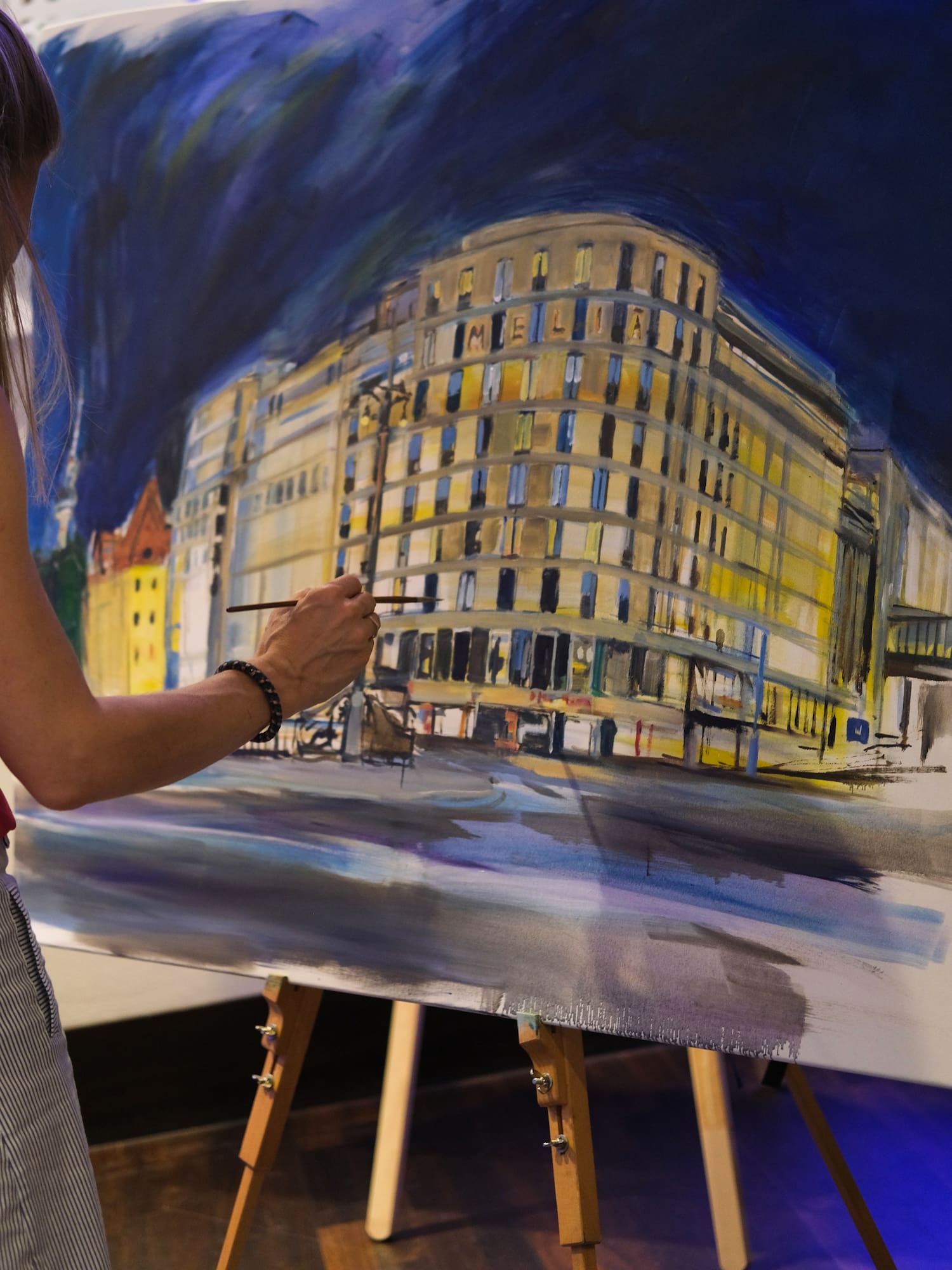 Artist's hand painting night cityscape with 'MELIA' building artwork.