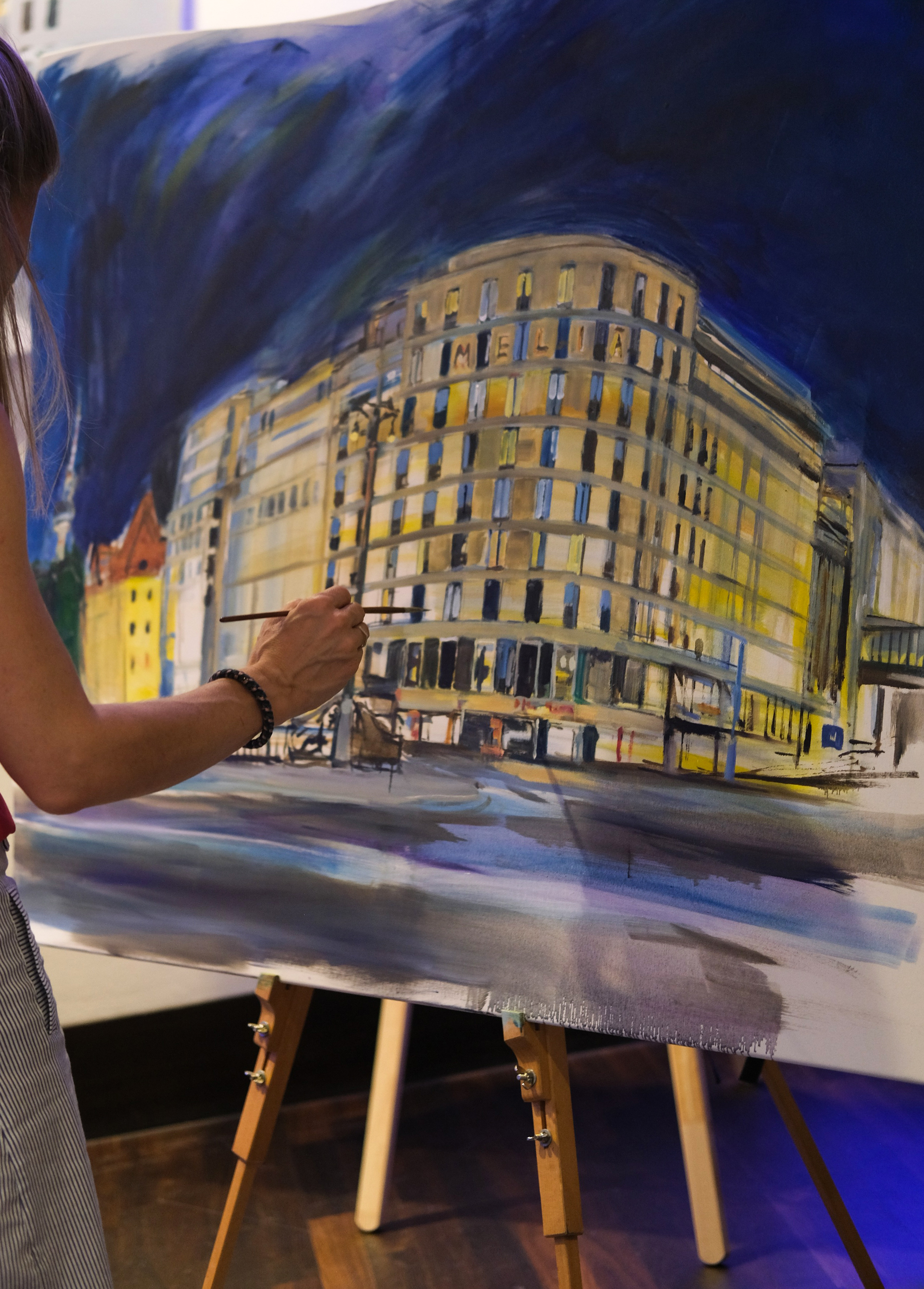 Artist's hand painting night cityscape with 'MELIA' building artwork.