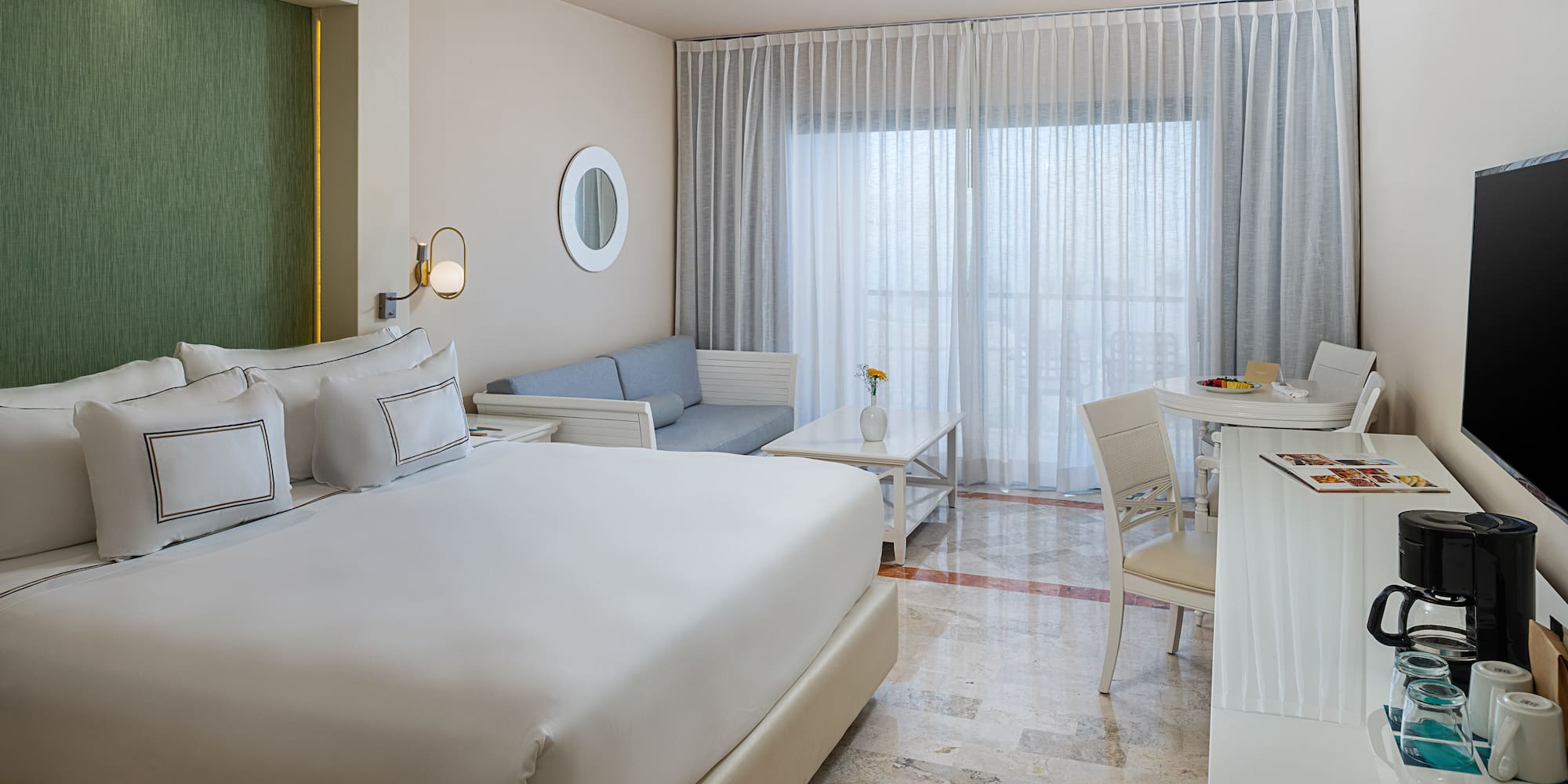Spacious modern hotel room with king bed, sofa, and dining area by bright window.
