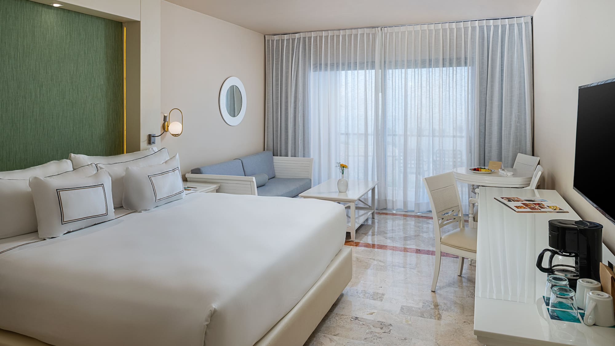 Spacious modern hotel room with king bed, sofa, and dining area by bright window.